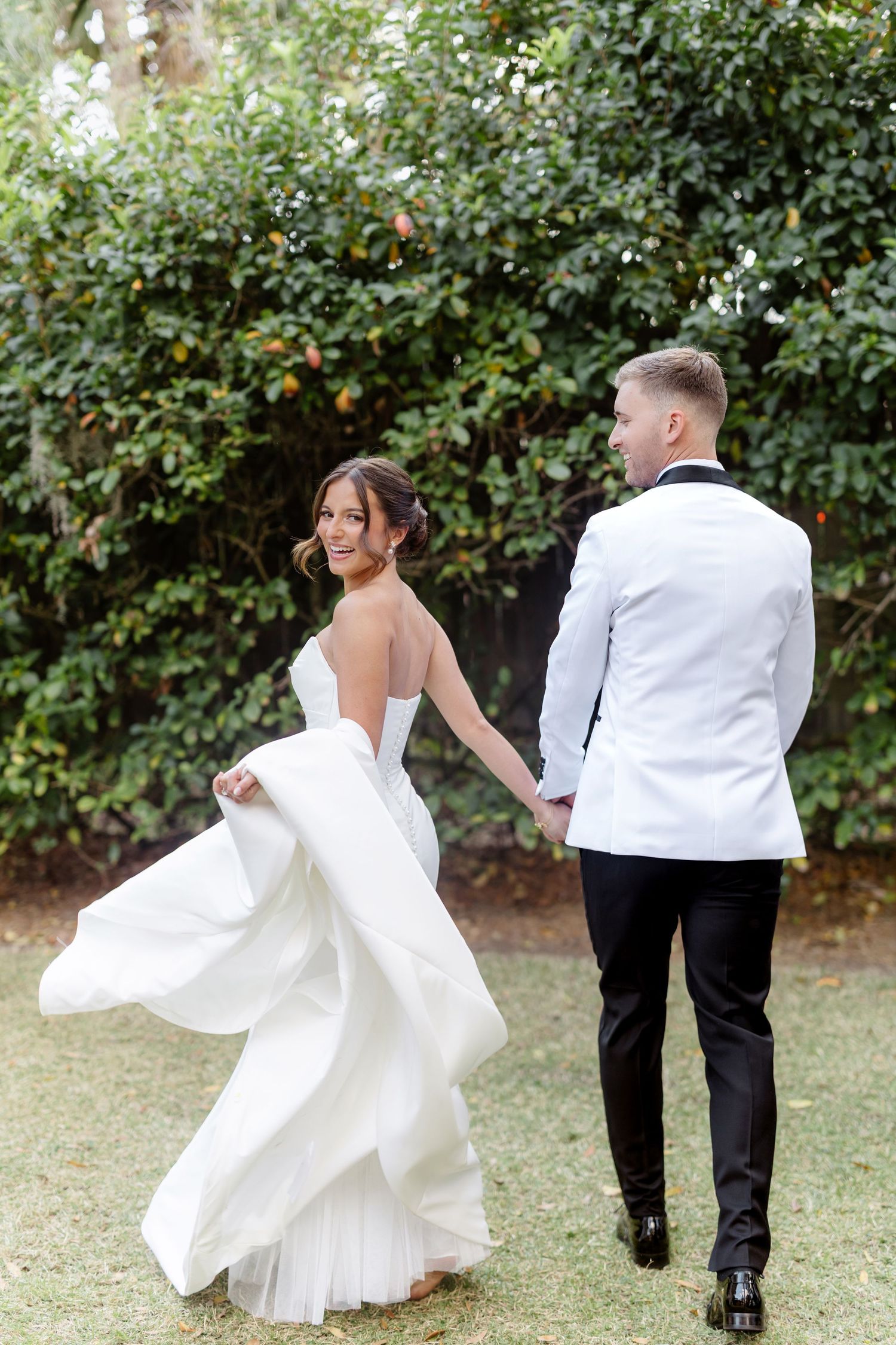 A couple in wedding attire playfully dances together in a garden setting with lush green foliage in the background.