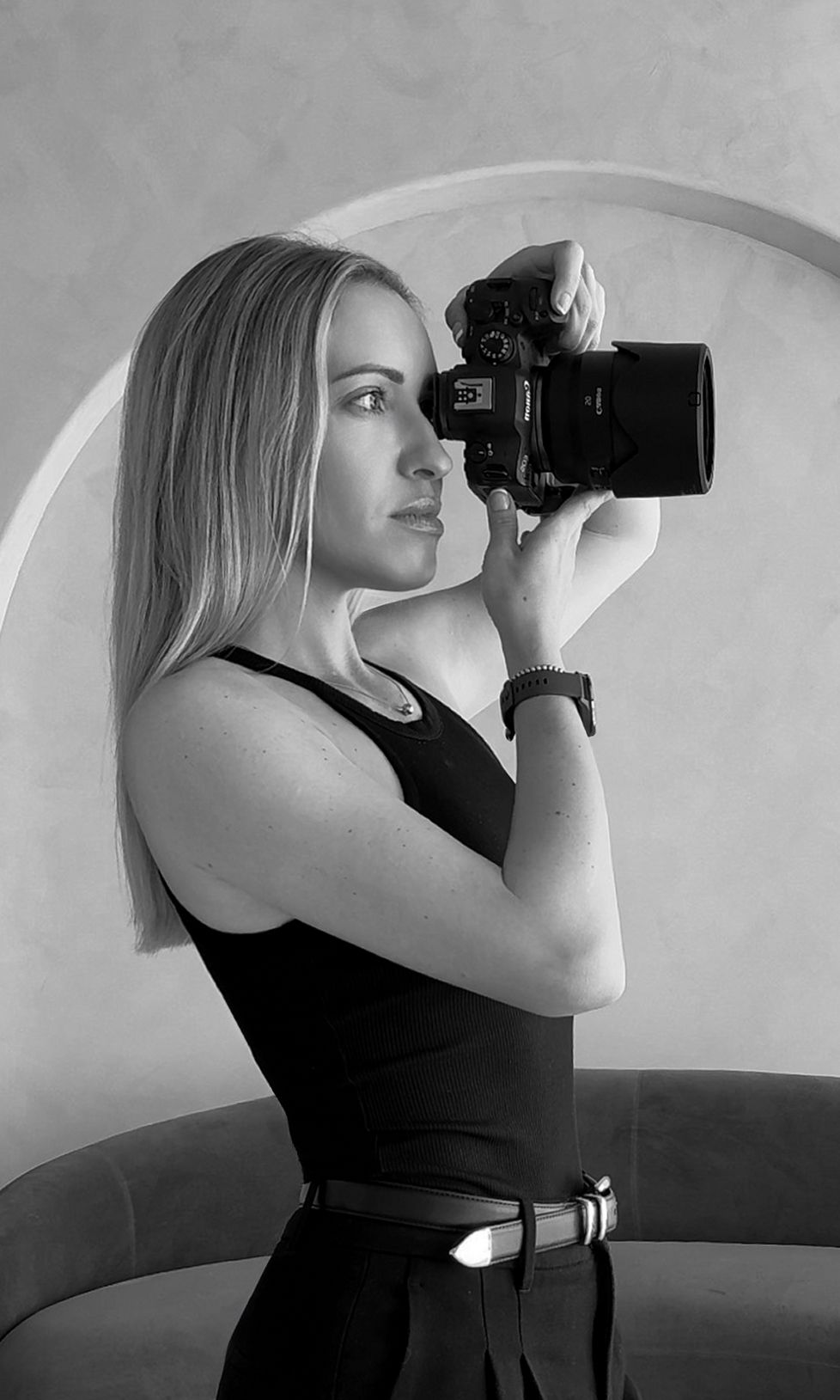Black and white photo of a blonde woman in a black dress holding a DSLR camera up to her eye.