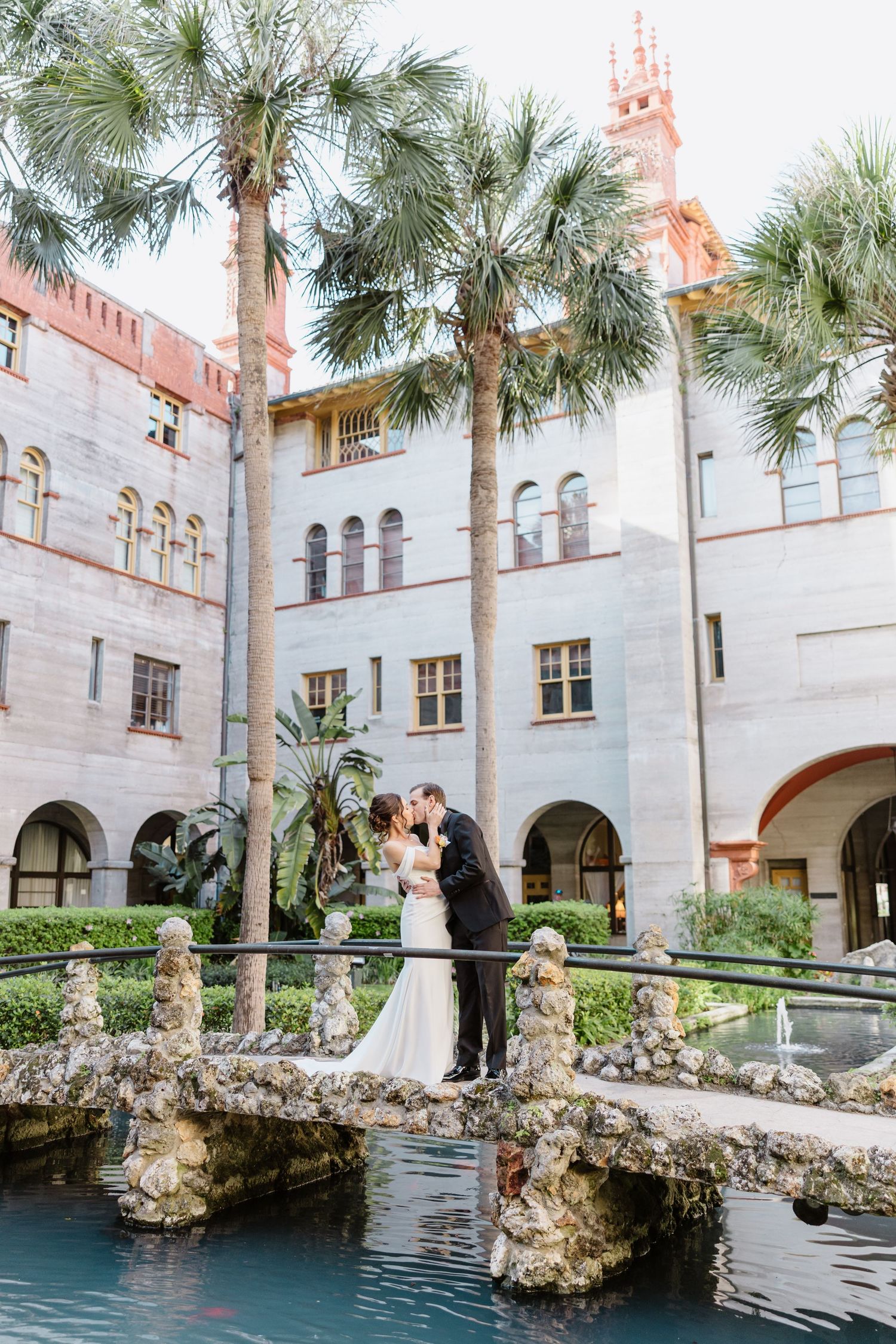 Wedding couple poses in front of historic Spanish Renaissance architecture with palm trees and waterfront views at dusk.