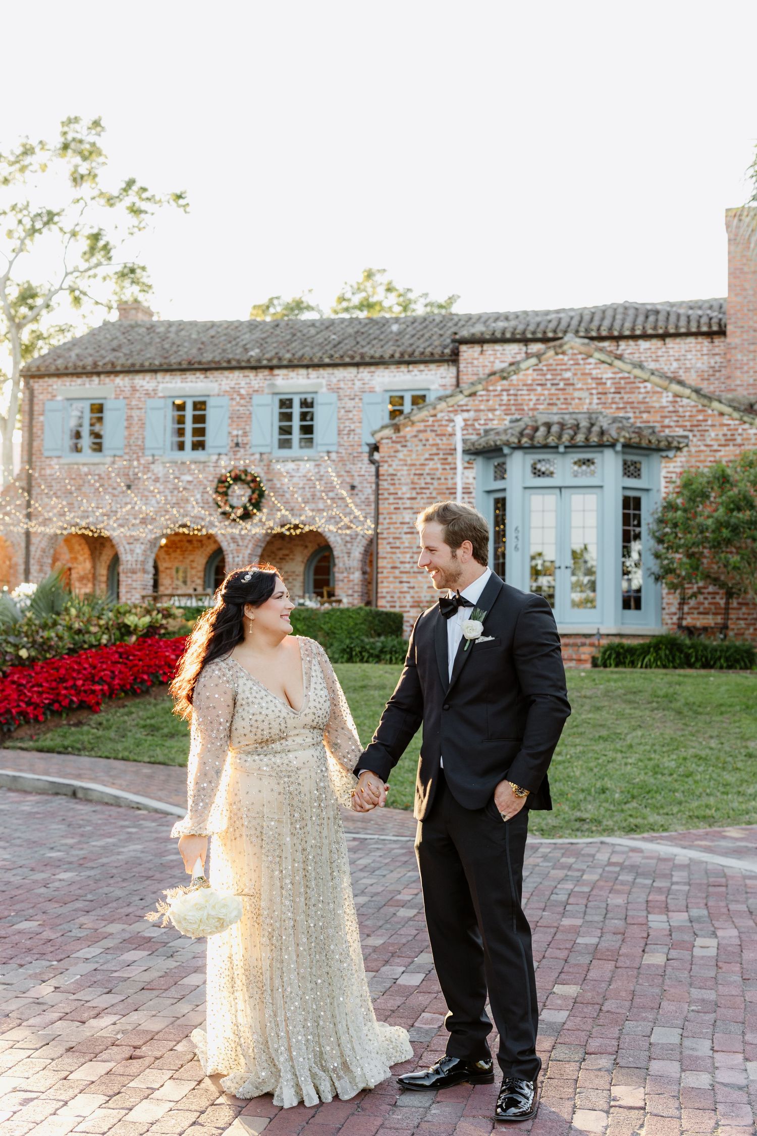 A couple in formal wedding attire walks along a brick pathway outside a historic building with blue windows.