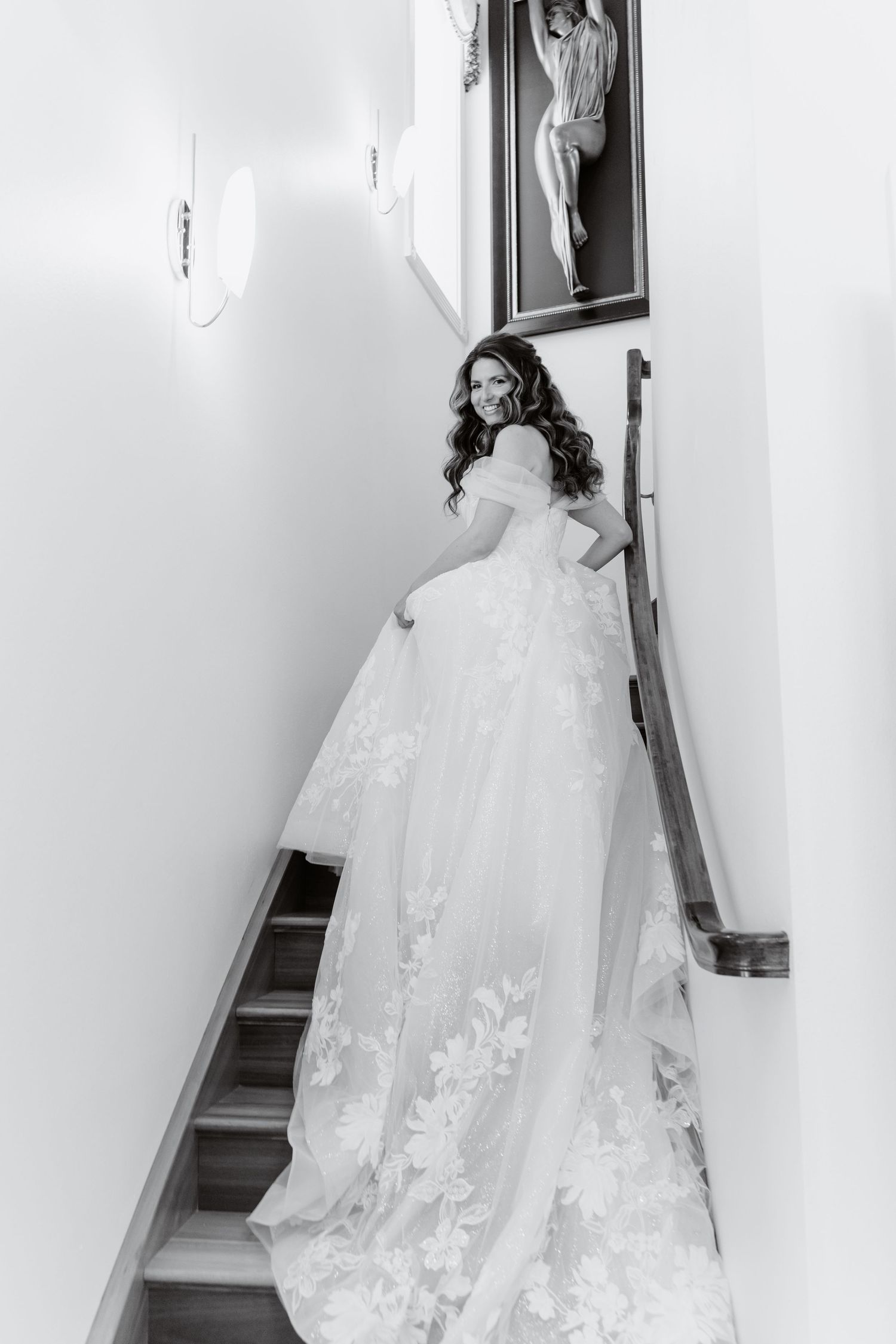 Bride in floral lace ball gown smiling on staircase, black and white wedding portrait photography.