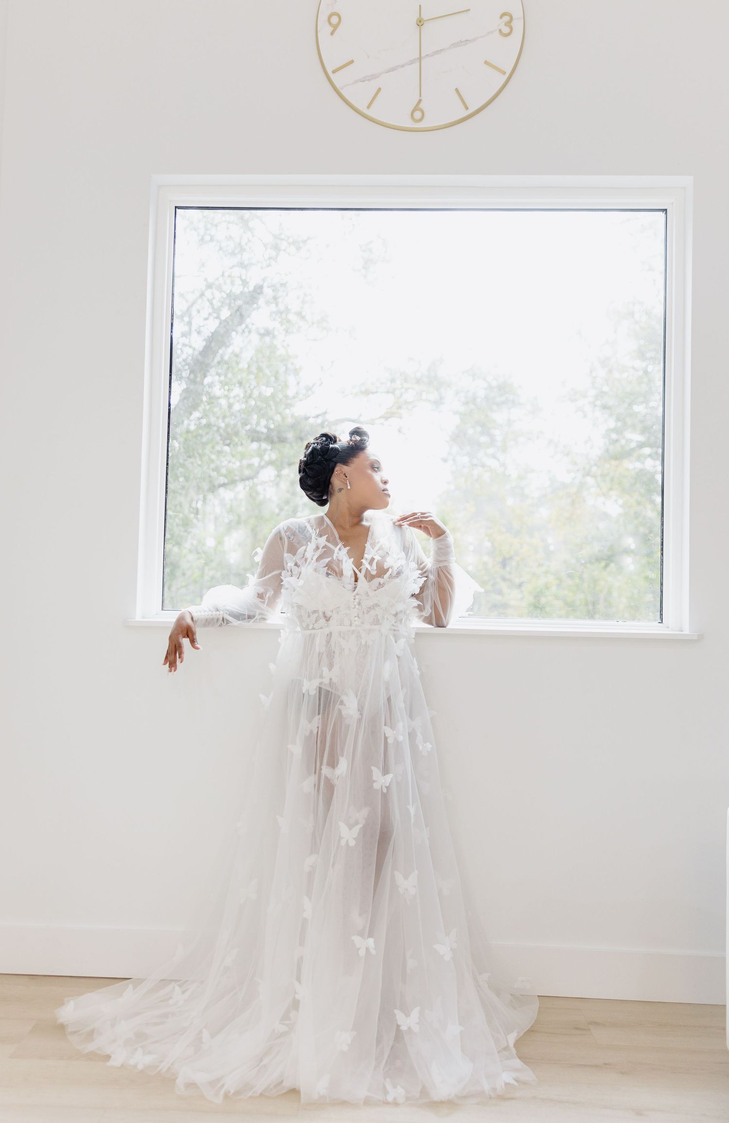 Bride in sheer white butterfly appliqué gown poses gracefully by a bright window with a gold clock above.