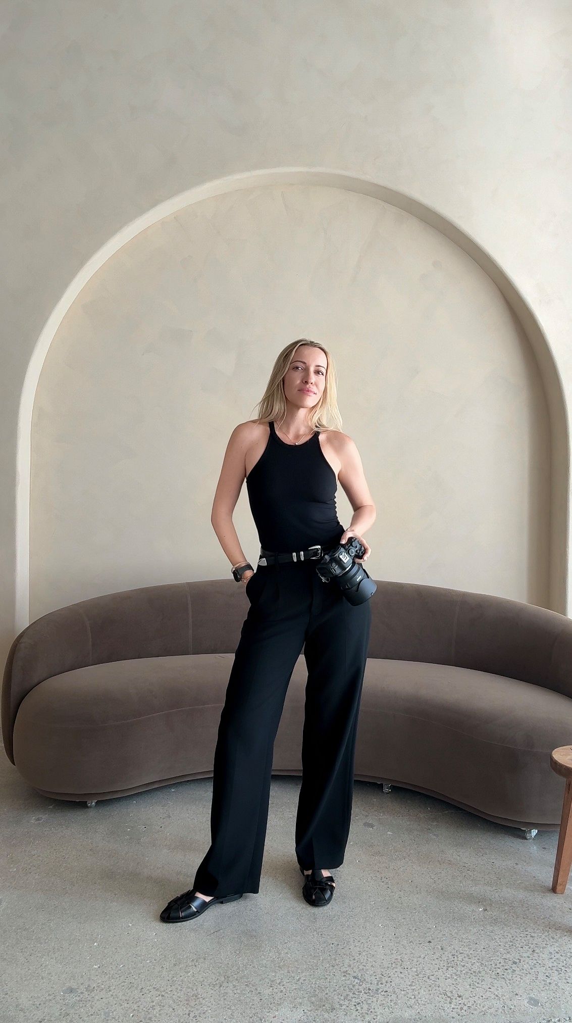 Woman in all-black outfit with halter top, wide-leg pants, and belt bag poses in minimalist interior with curved sofa.