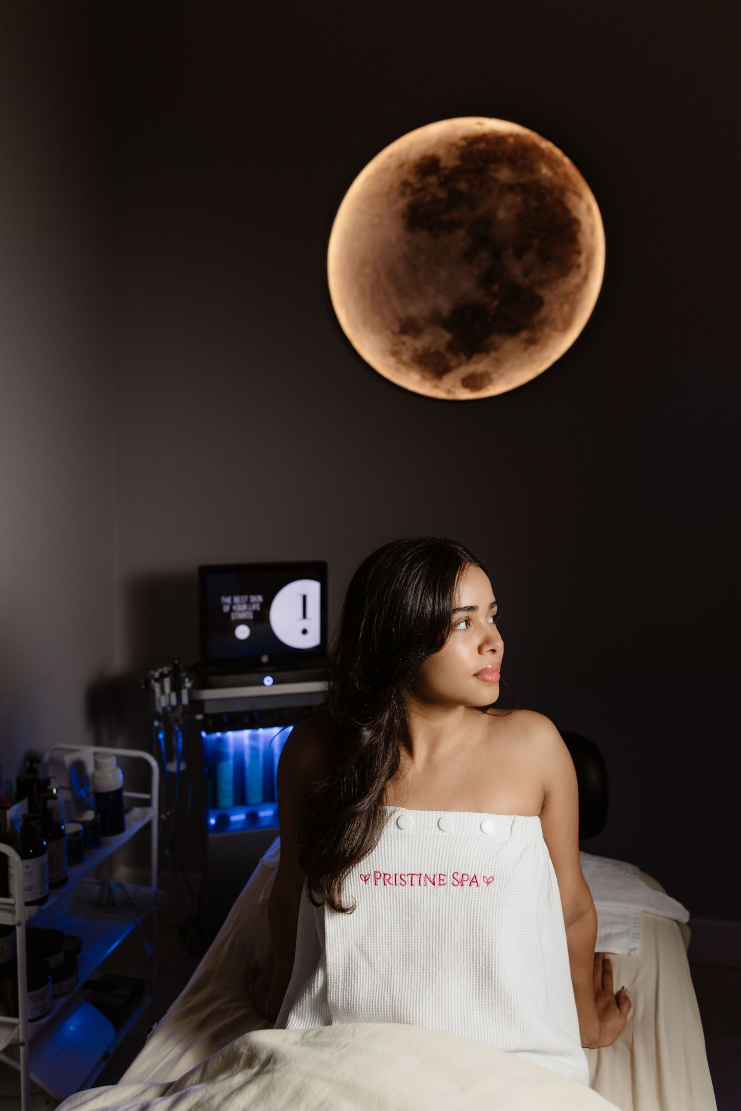 Woman wrapped in Pristine Spa towel sitting in dimly lit spa room with glowing moon wall decor and blue-lit equipment.