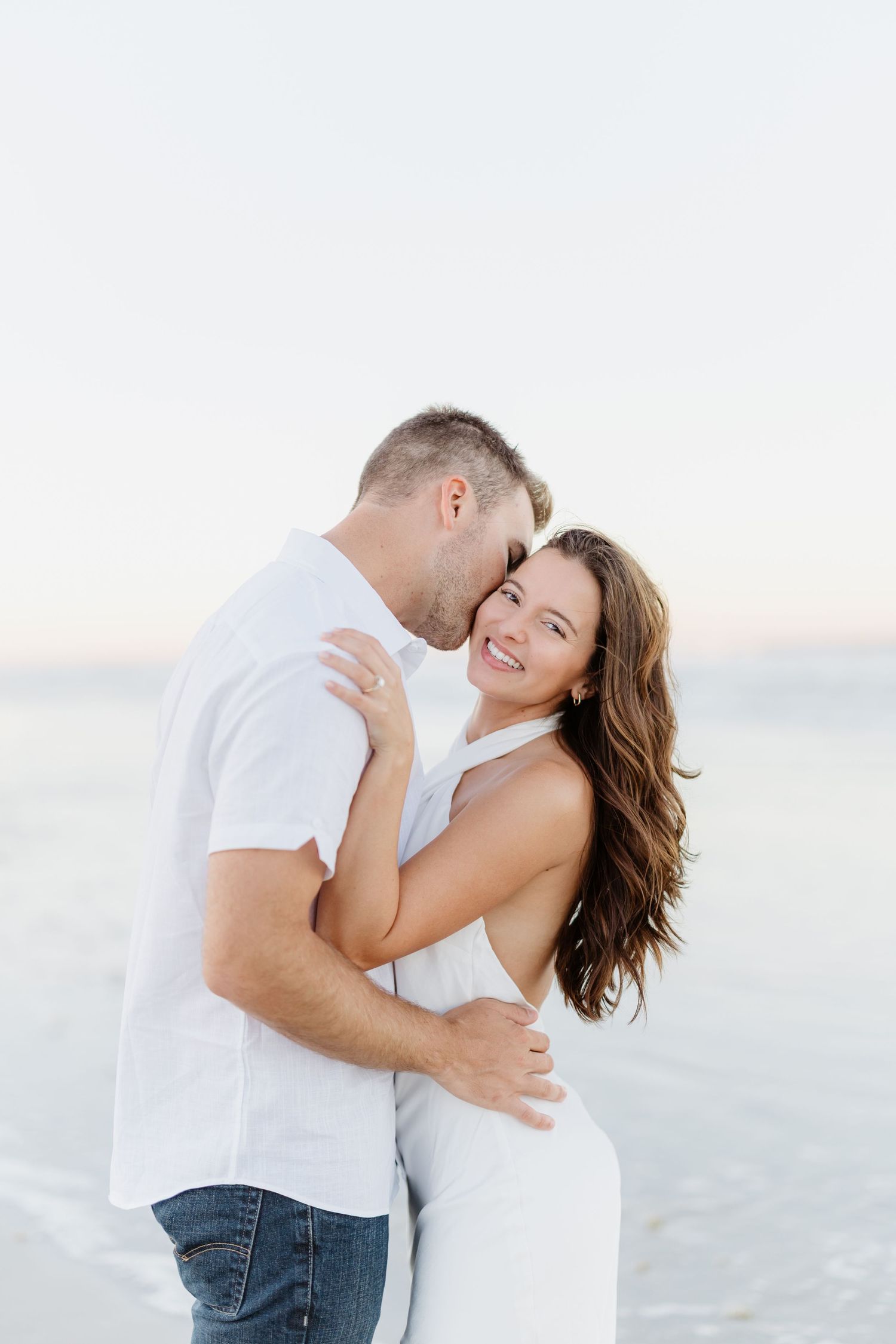 A romantic couple in white outfits embraces and laughs together during a beach photoshoot at sunset.
