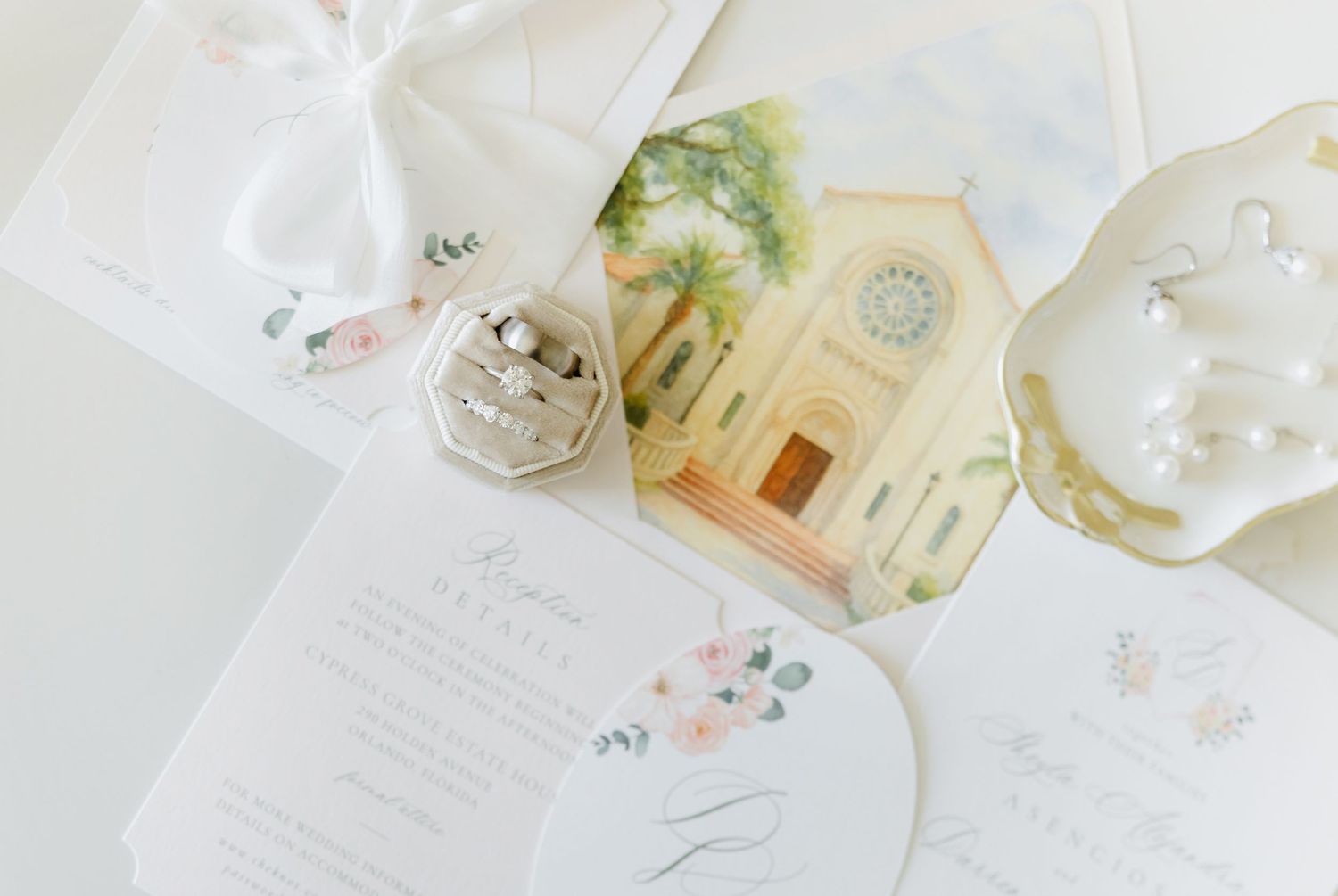 Elegant wedding flat lay with floral invitations, watercolor church illustration, diamond rings, and pearl earrings on white background.