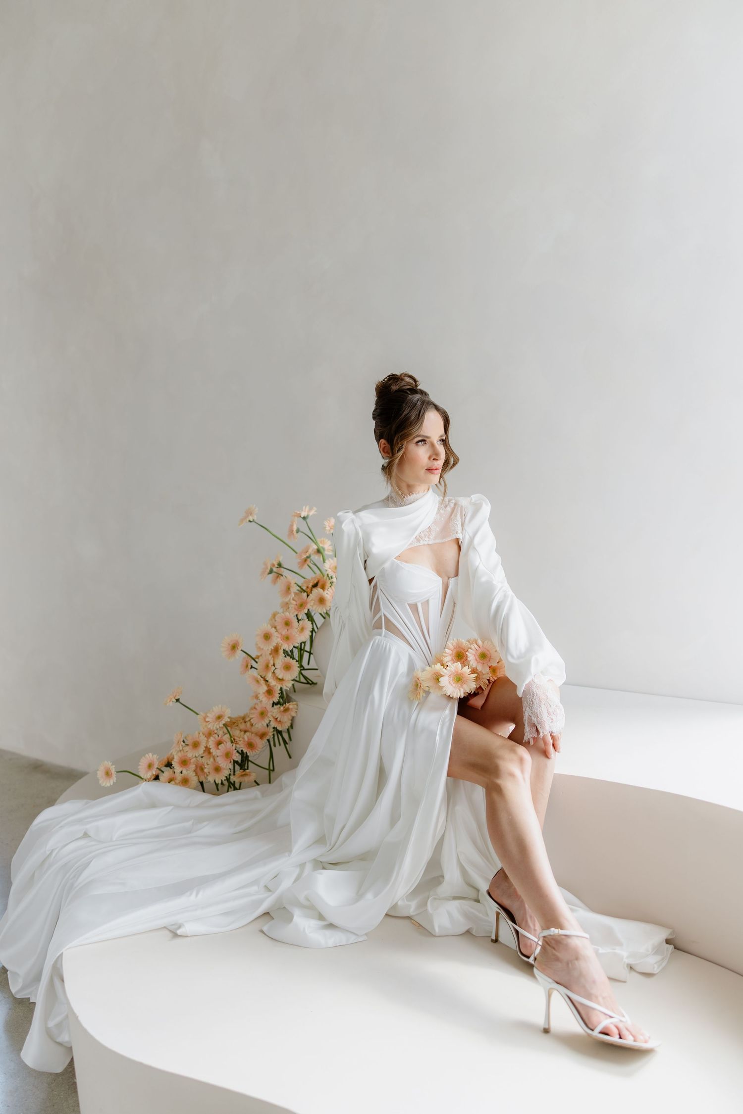 Elegant bride in flowing white robe gown seated with peach flowers, wearing strappy heels in a minimalist studio setting.