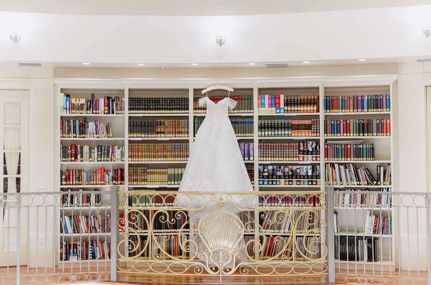 White wedding dress hanging on ornate railing in front of floor-to-ceiling bookshelves filled with colorful books.