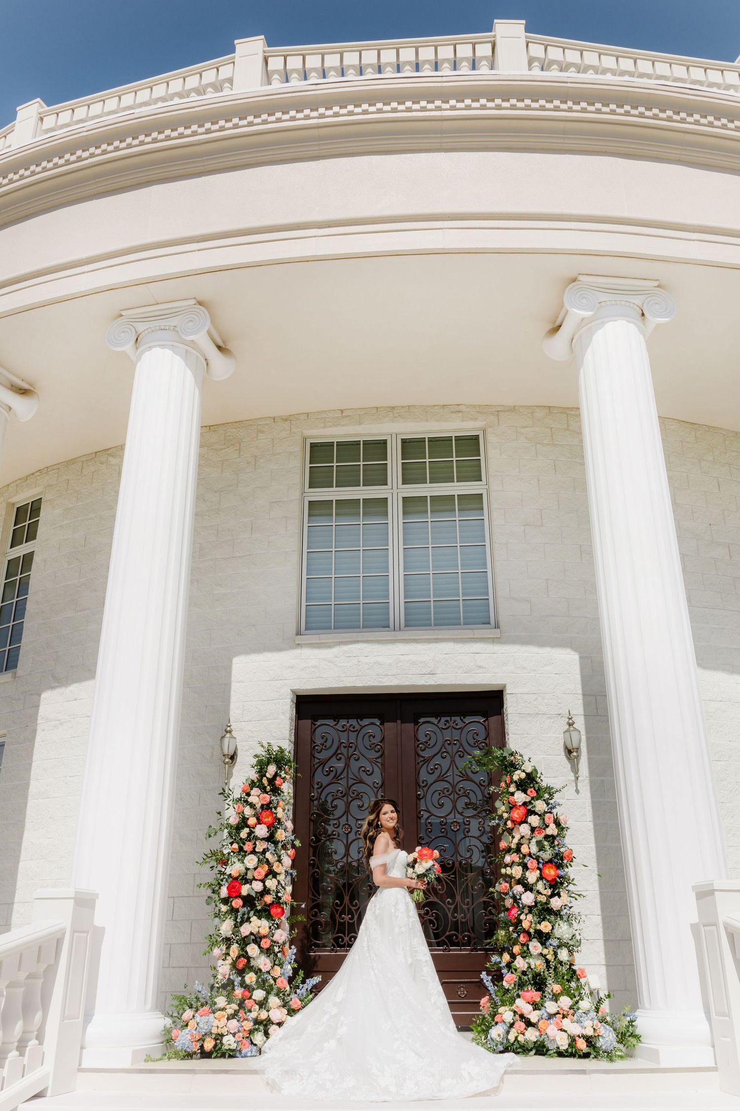 Bride in white gown holding bouquet stands between floral arches at elegant venue with white columns.