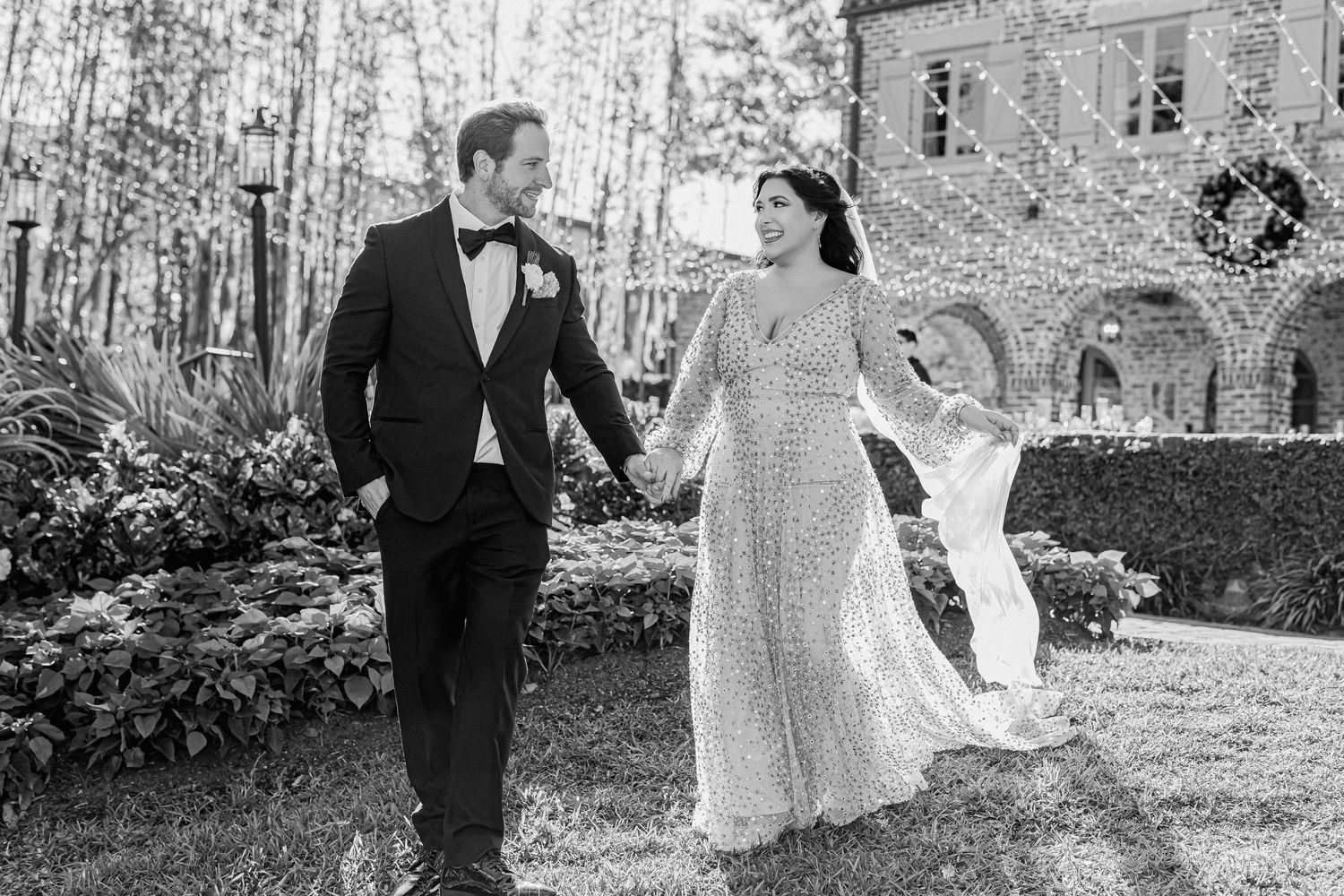Wedding couple walks hand in hand with flowing dress in black and white outdoor setting.