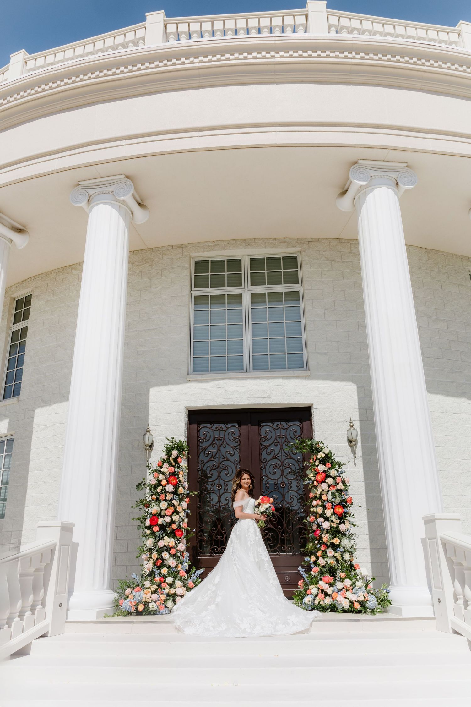 Bride in white gown stands on steps of elegant venue with floral arches and tall white columns.