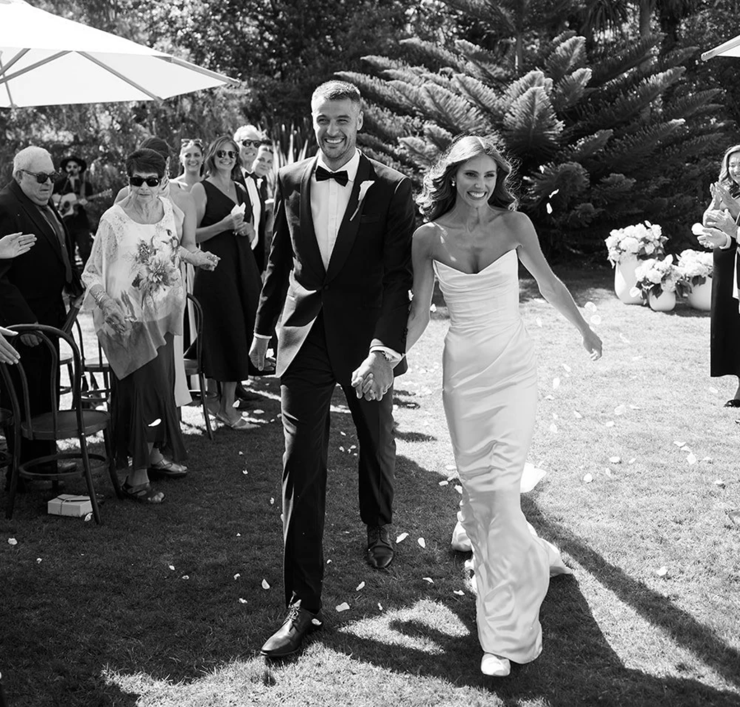 Black and white photo of a smiling bride and groom walking hand in hand down the aisle at an outdoor wedding ceremony.