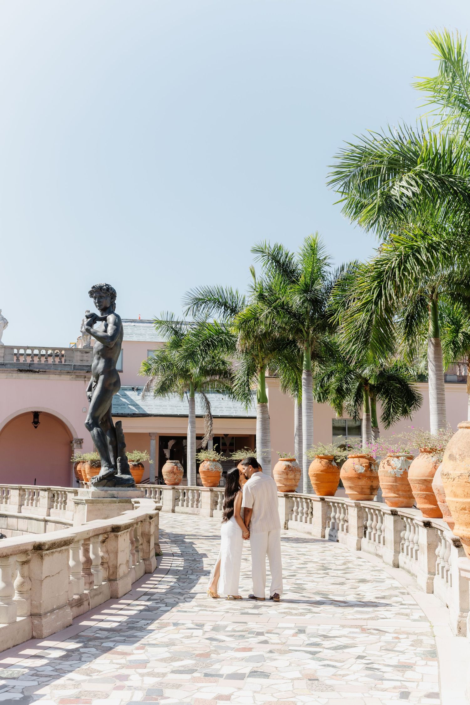 Couple in white attire strolling past a David statue and terracotta urns on a sunlit Mediterranean-style terrace with palm trees.
