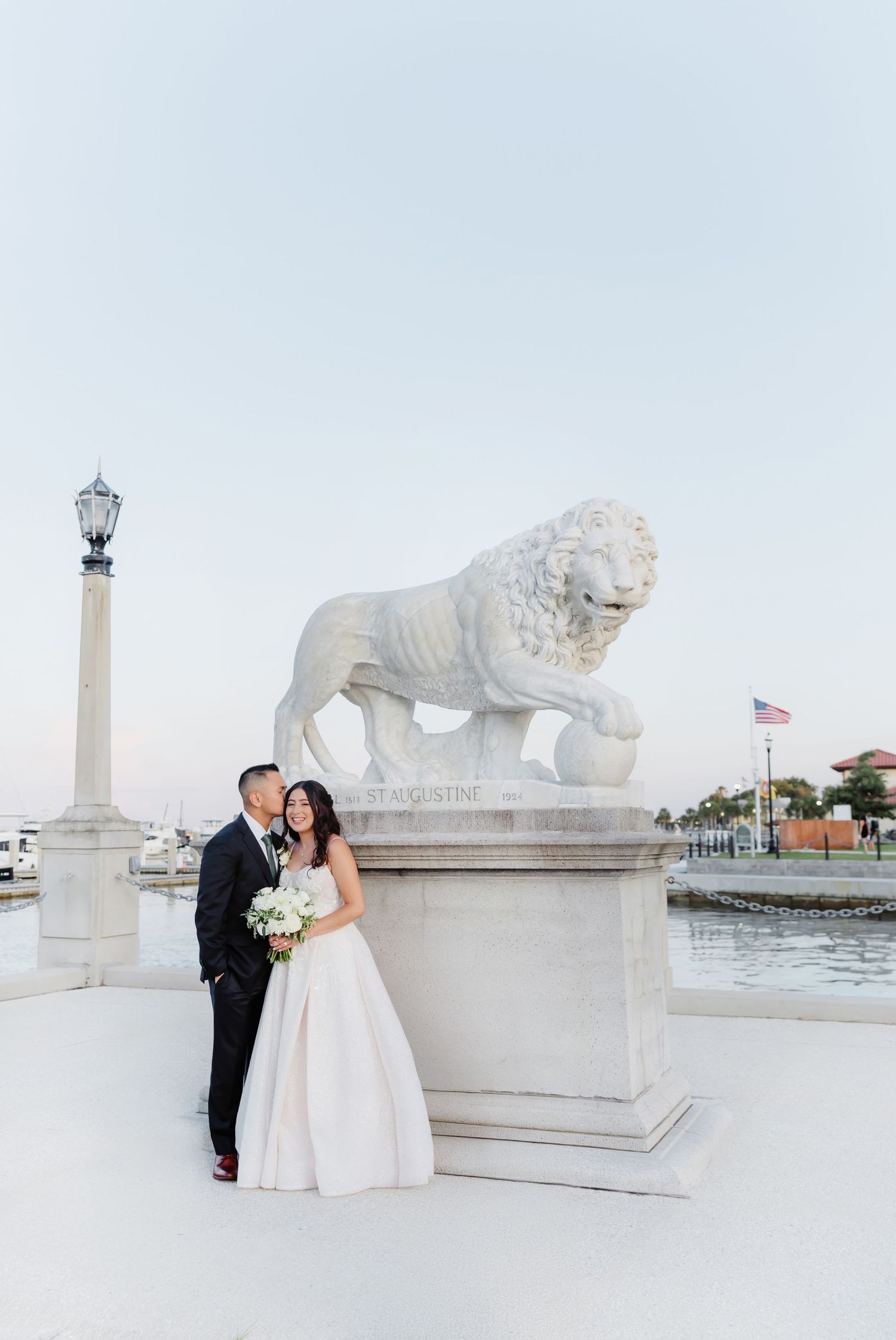 A couple shares romantic moments at a grand outdoor venue with ornate stone lion statues and columns at sunset.