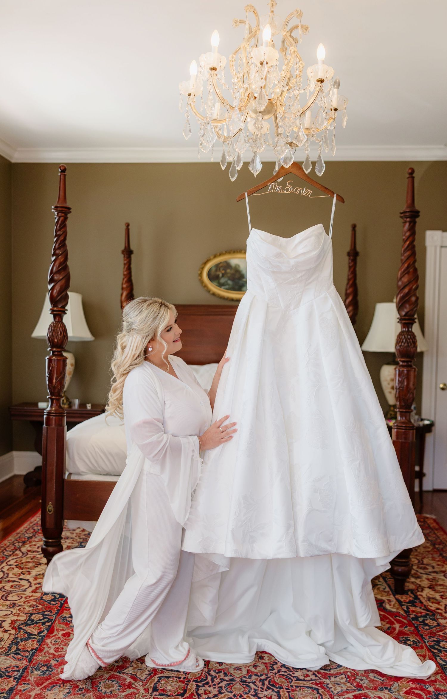 Wedding inspo Bride in white robe admiring her strapless ball gown wedding dress hanging from a chandelier in an elegant bedroom.