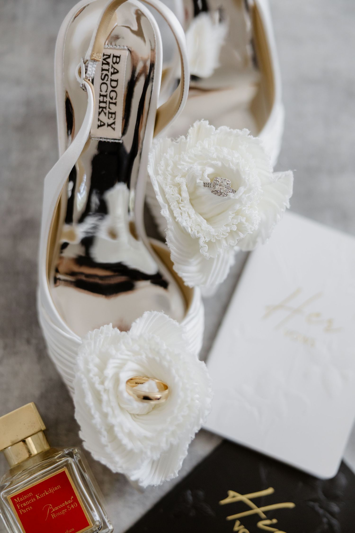 White Badgley Mischka bridal heels with floral detail, gold wedding rings, Maison Francis Kurkdjian perfume, and vow book.