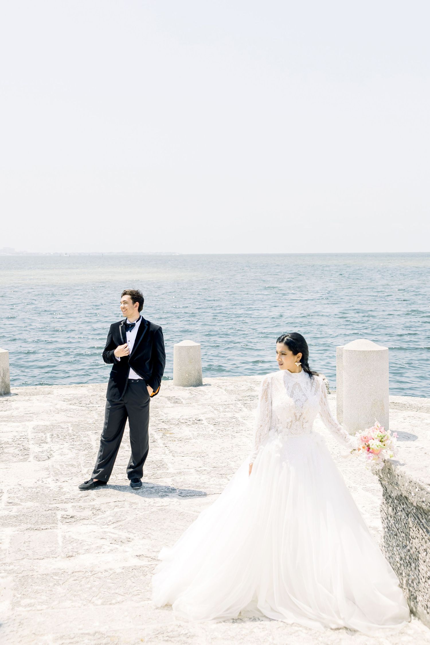 Elegant wedding couple poses along an oceanfront stone wall in formal attire with coastal views on a bright sunny day.