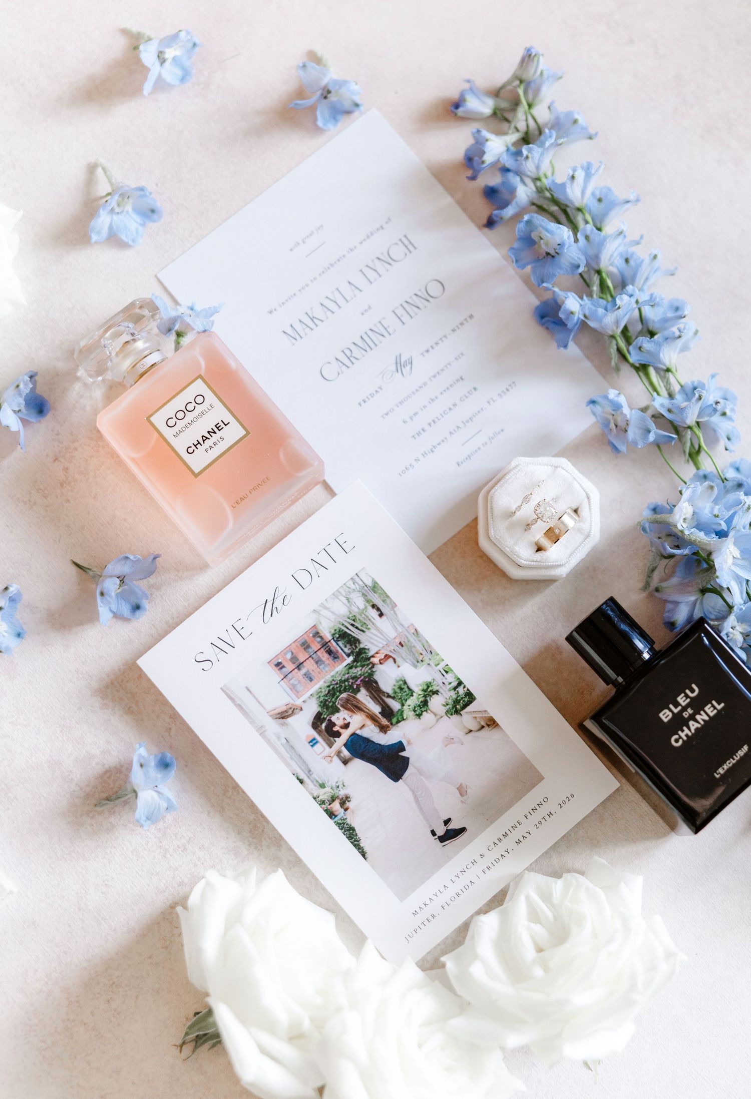 Elegant wedding flat lay with Chanel perfume, save the date card, floral invitation, ring box, and blue flowers.