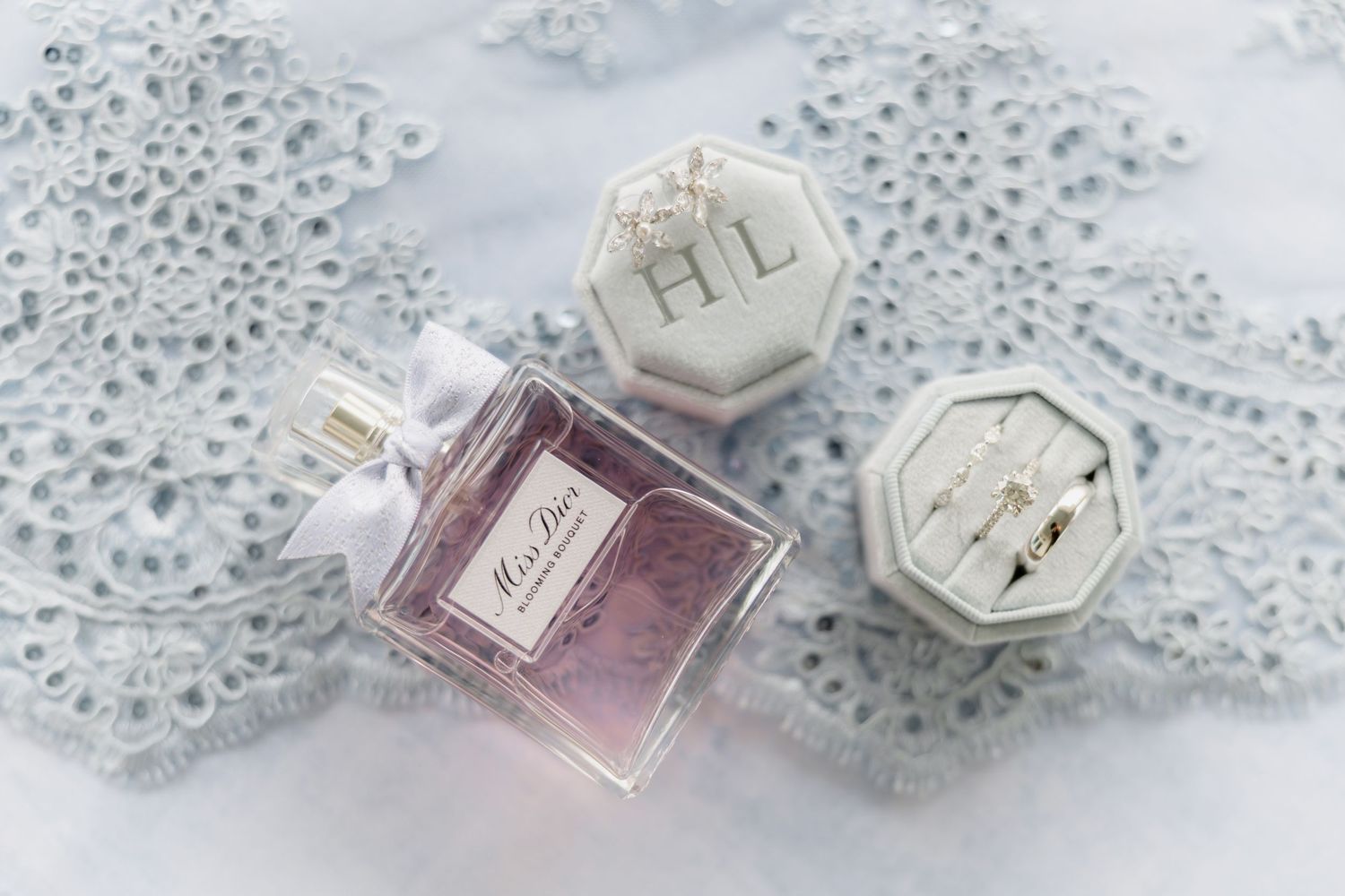 Miss Dior perfume bottle beside a hexagonal ring box with wedding rings on delicate lace fabric.