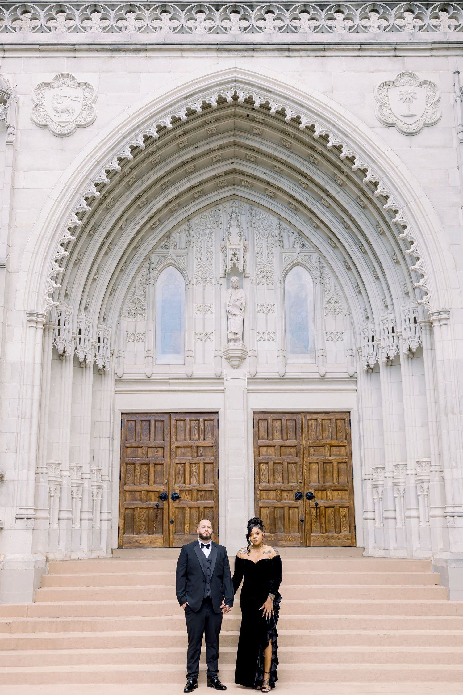 A couple stands in front of ornate Gothic church doors with beautiful white stone archways and steps at the entrance.