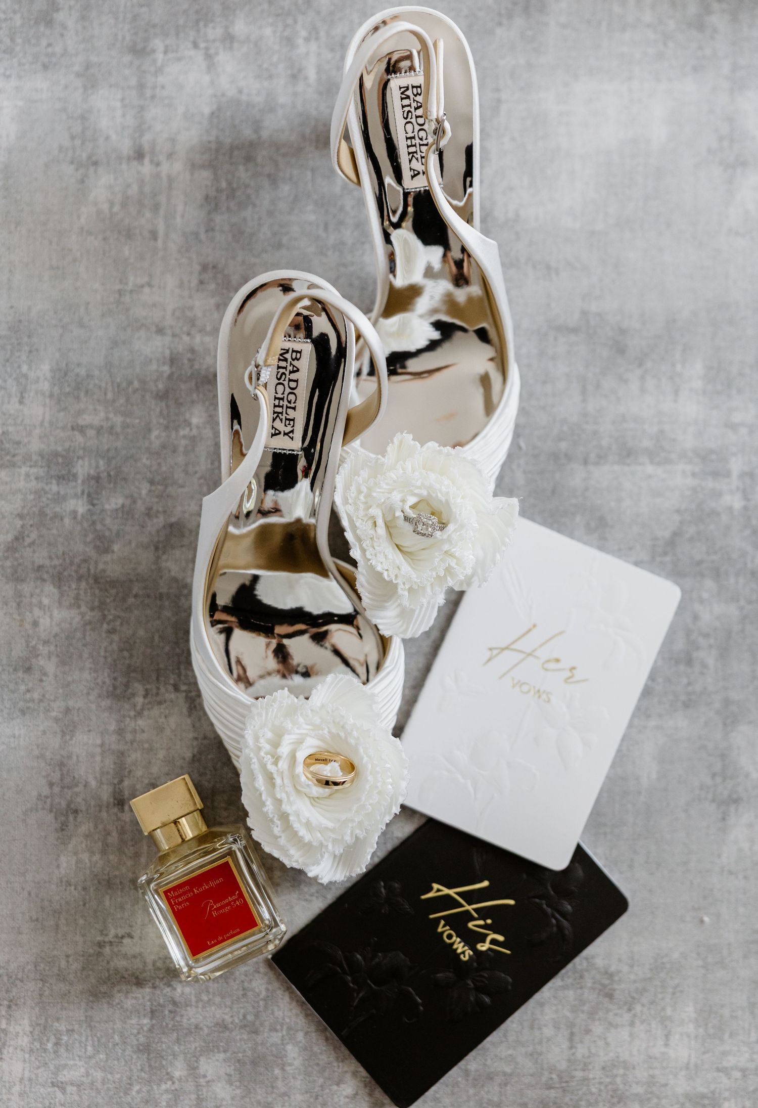 White Badgley Mischka bridal heels with floral detail, perfume bottle, and His and Hers vow books on gray background.