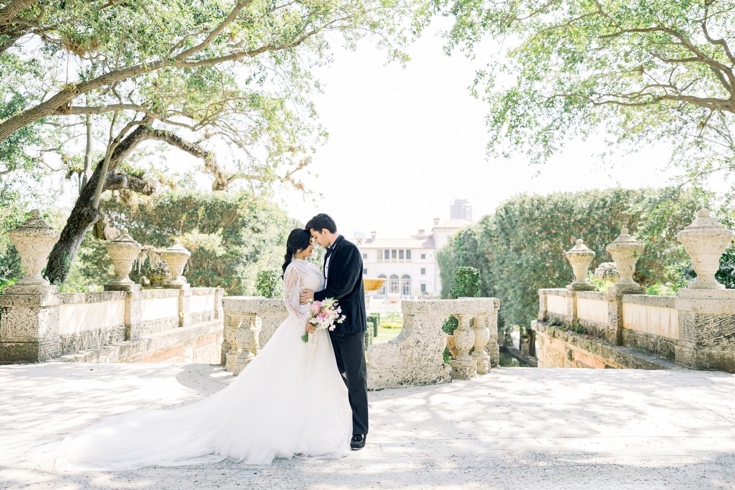 A couple embraces for a romantic wedding portrait under sunlit trees on a stone terrace.