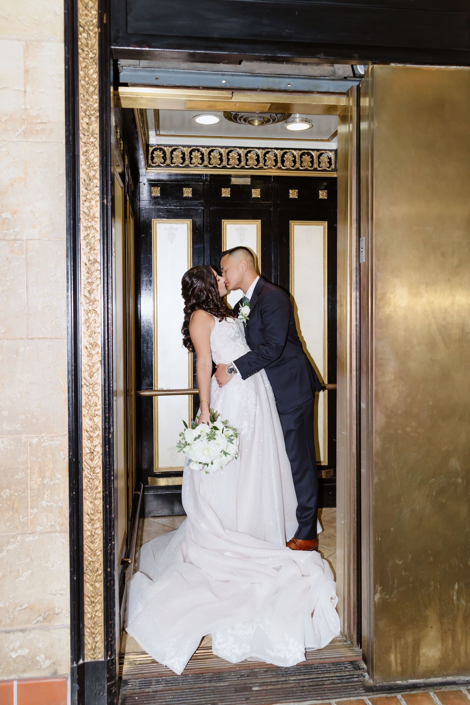 A romantic moment captured in an ornate gold elevator as a couple in wedding attire share a kiss while holding white flowers.