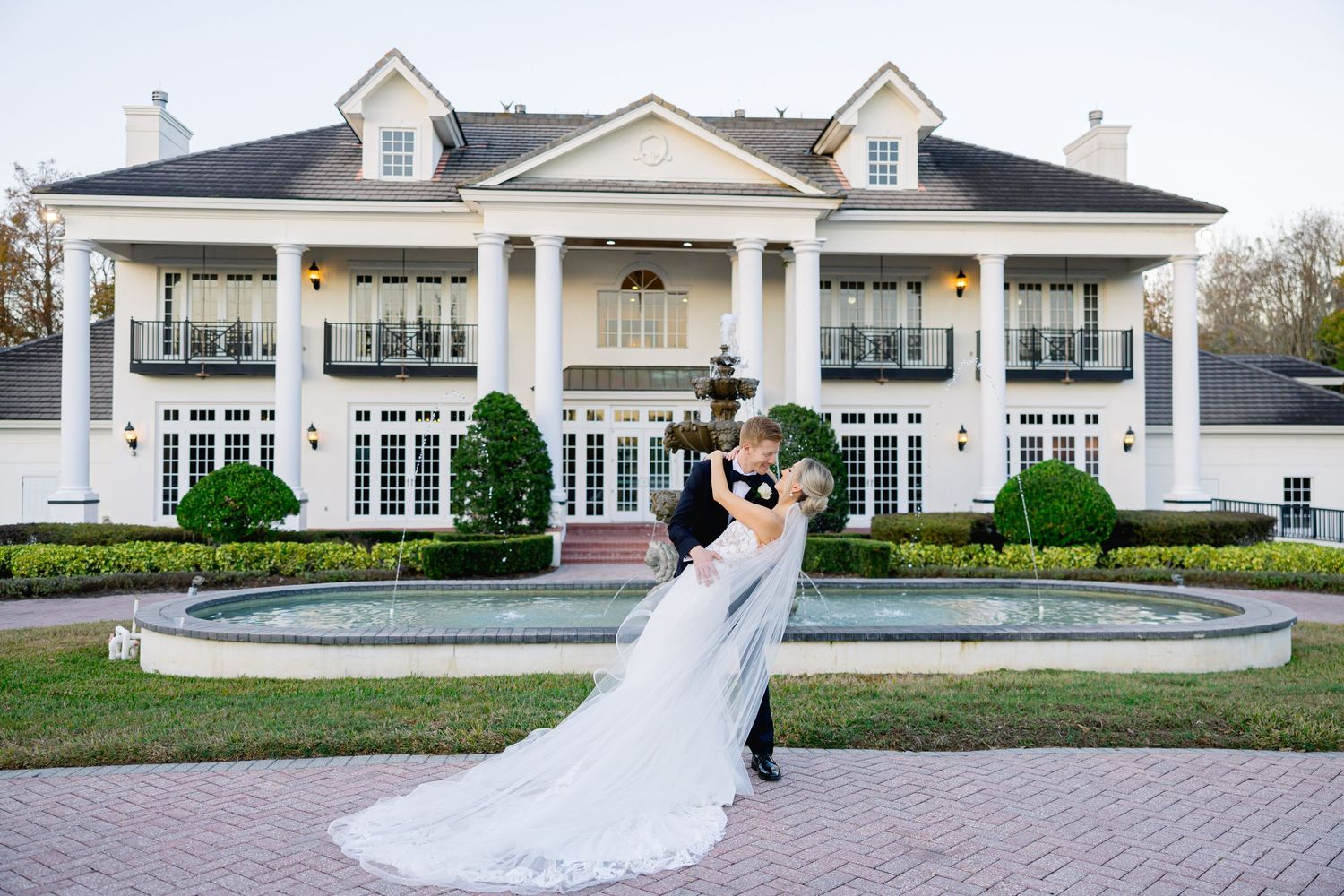 A bride and groom embrace in front of a grand white colonial mansion with columns.
