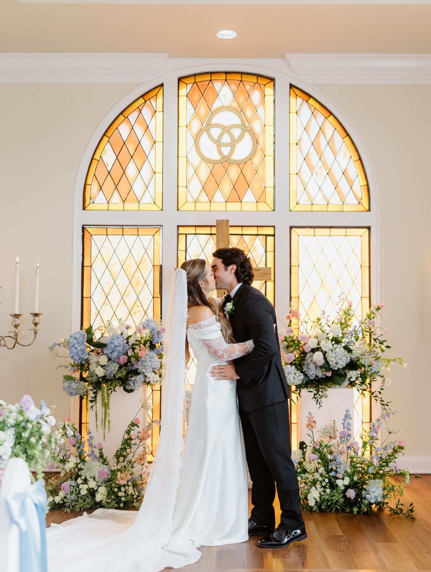 Bride and groom share first kiss at altar with stained glass window and pastel floral arrangements in elegant chapel.