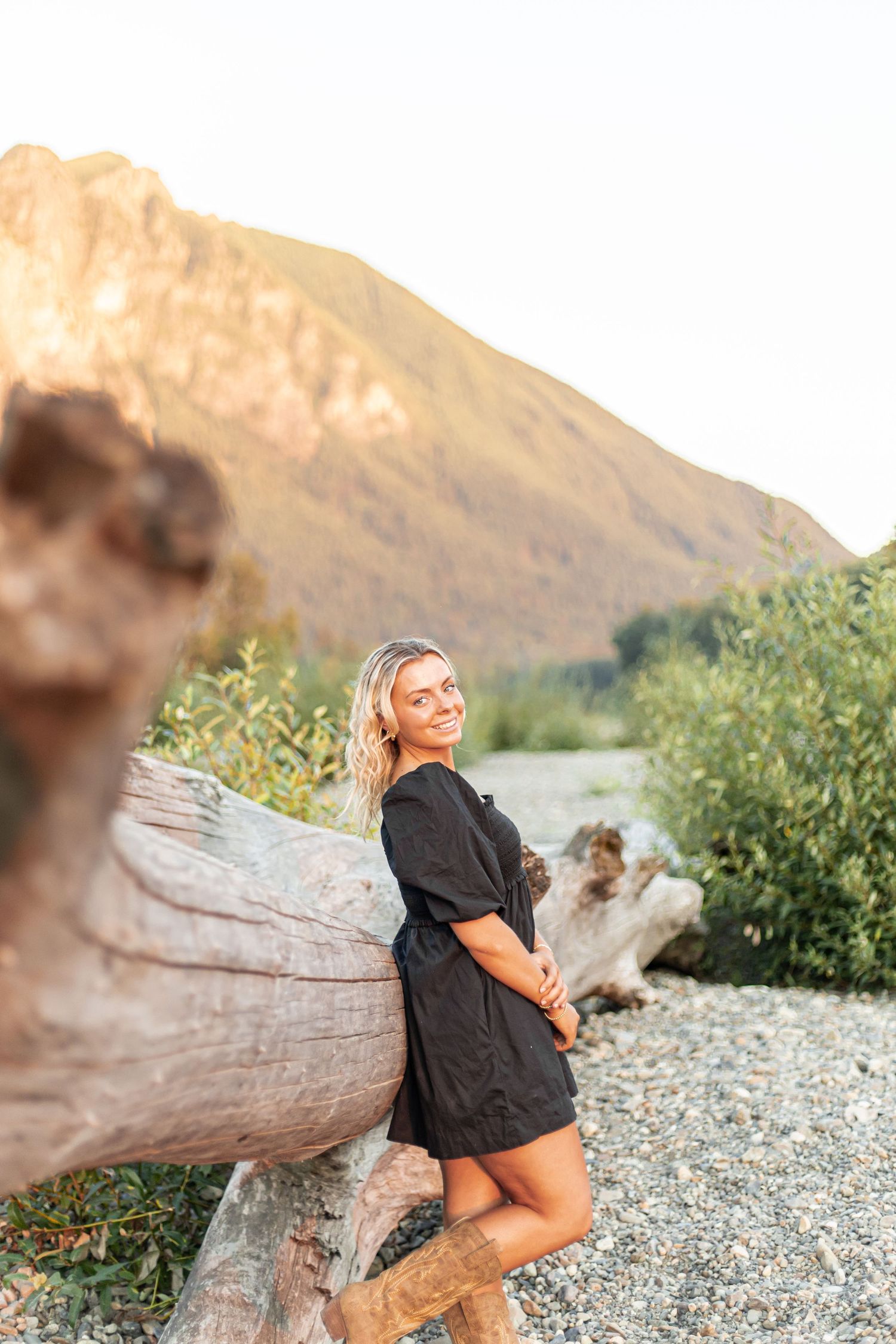 A person sits on a large driftwood log at a scenic riverside with mountains in the background during golden hour.