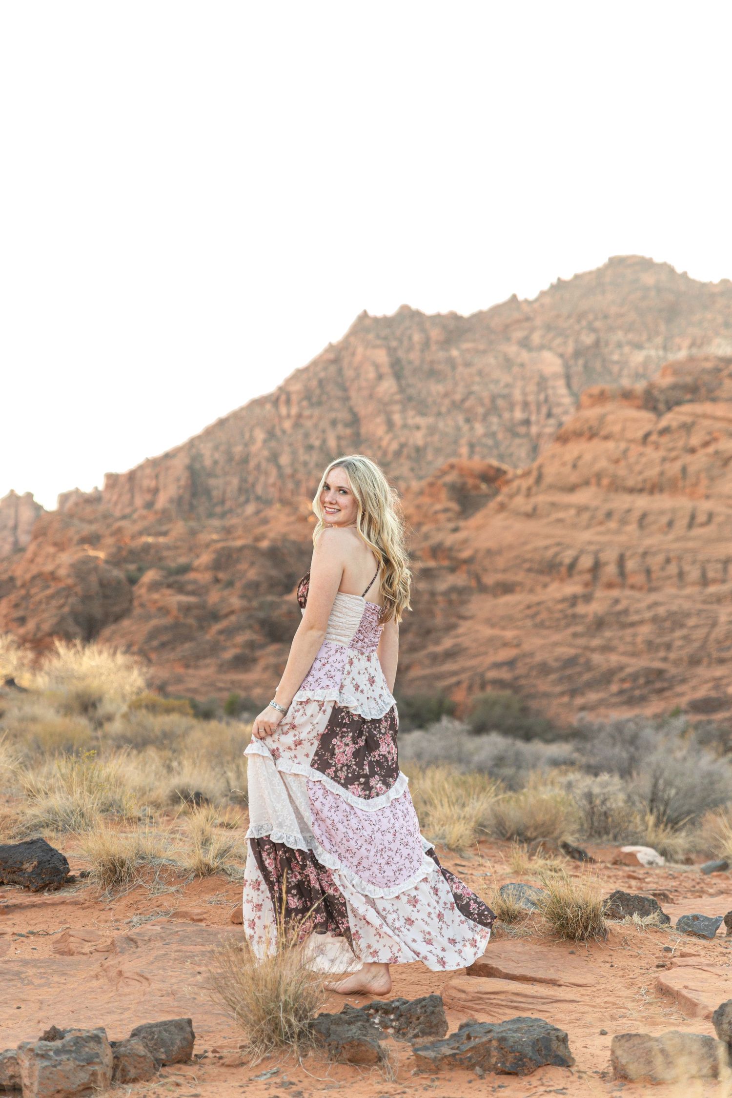 A person in a flowing dress poses gracefully on red rocks in a desert landscape at sunset.