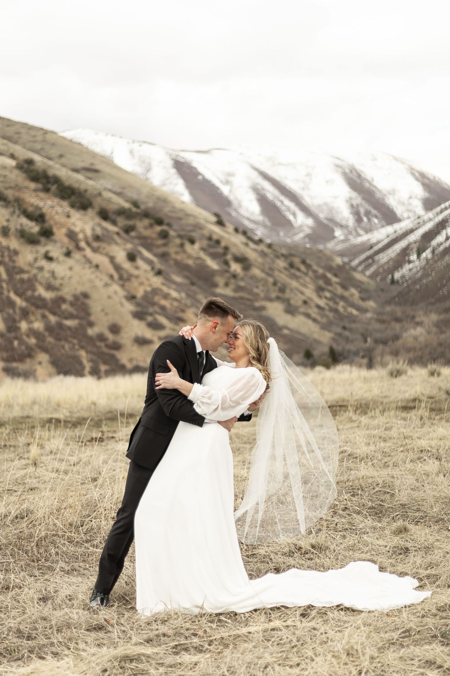 A couple in wedding attire embraces on a grassy hillside with snow-capped mountains in the background.