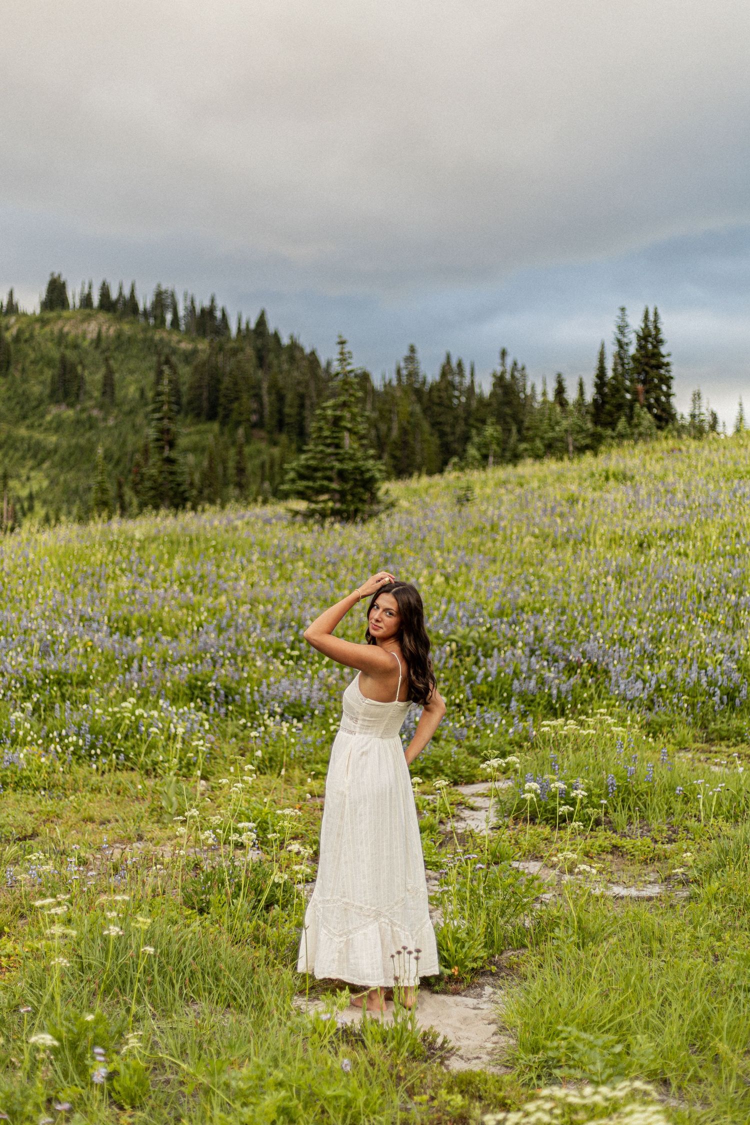 A white sundress twirls through a mountain meadow filled with purple wildflowers and evergreen trees under cloudy skies.