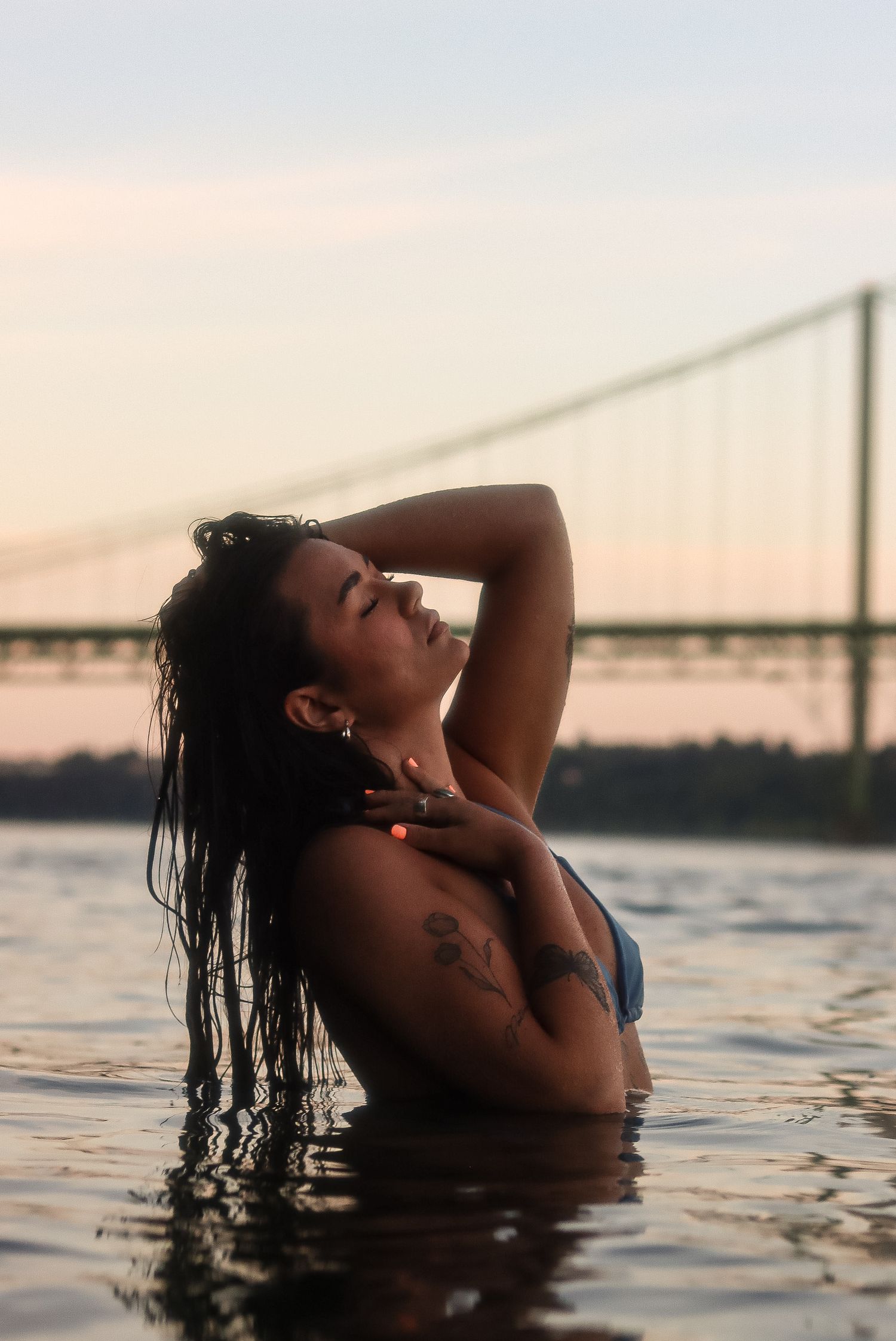Cora Emilie Media A silhouetted figure bathes in calm waters at sunset with a bridge visible in the background.