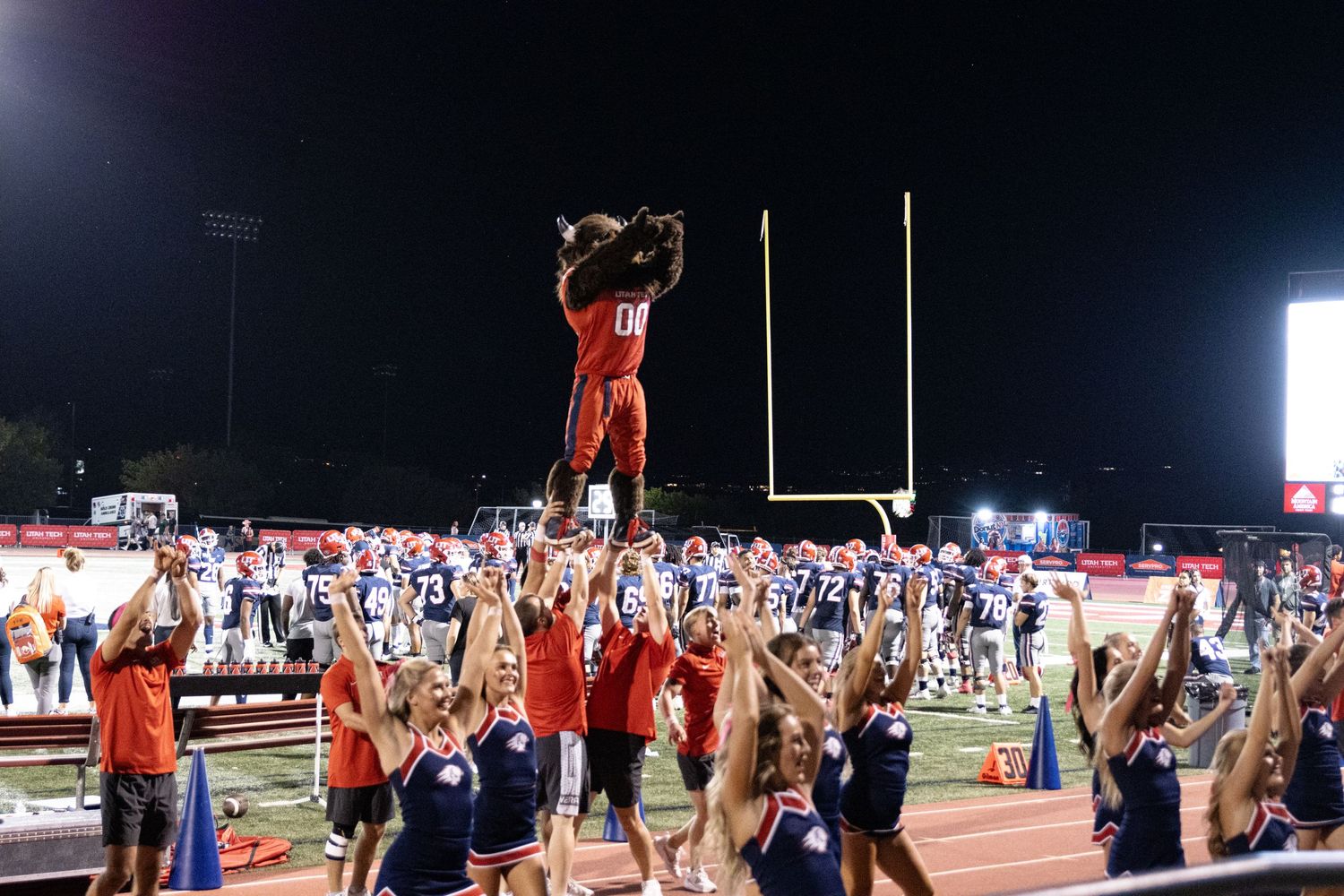 Cora Emilie Media Cheerleaders perform stunt lift during nighttime football game with bright stadium lights.