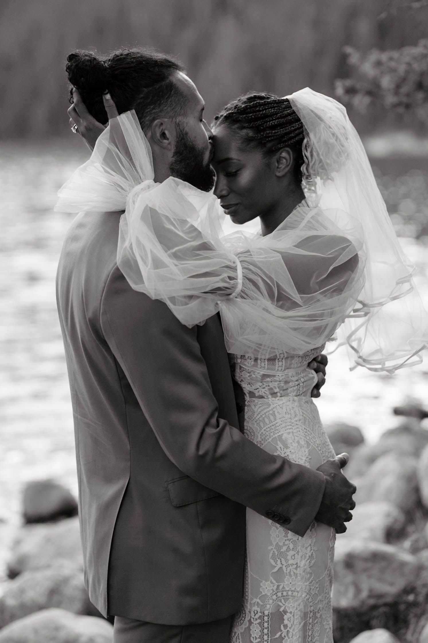 A romantic black and white wedding portrait shows a tender moment between newlyweds as the bride's veil flows in the breeze.