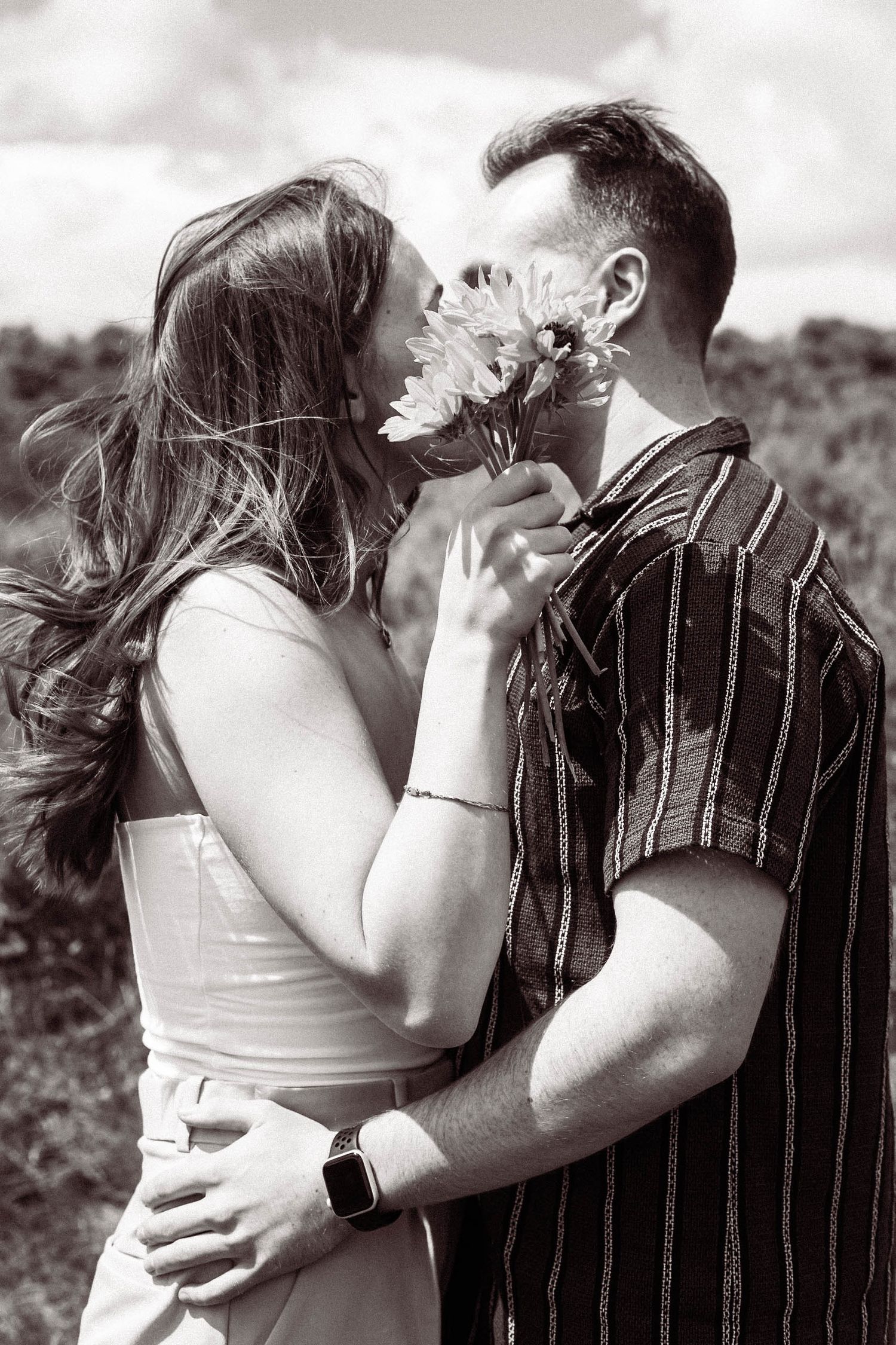A romantic black and white photo of a couple sharing a kiss behind a daisy flower in an outdoor setting.