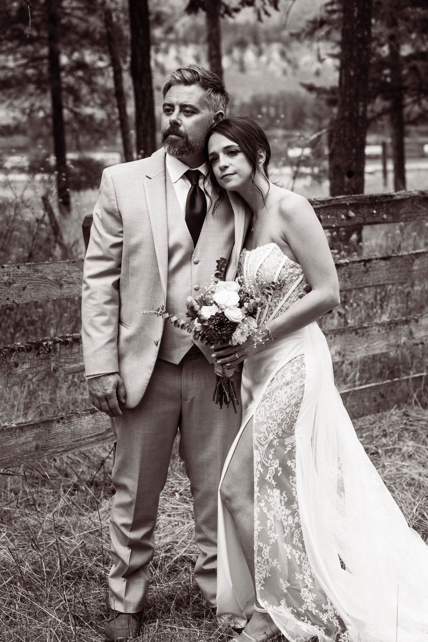 A romantic black and white photo of a couple in wedding attire posing outdoors near a rustic wooden fence.