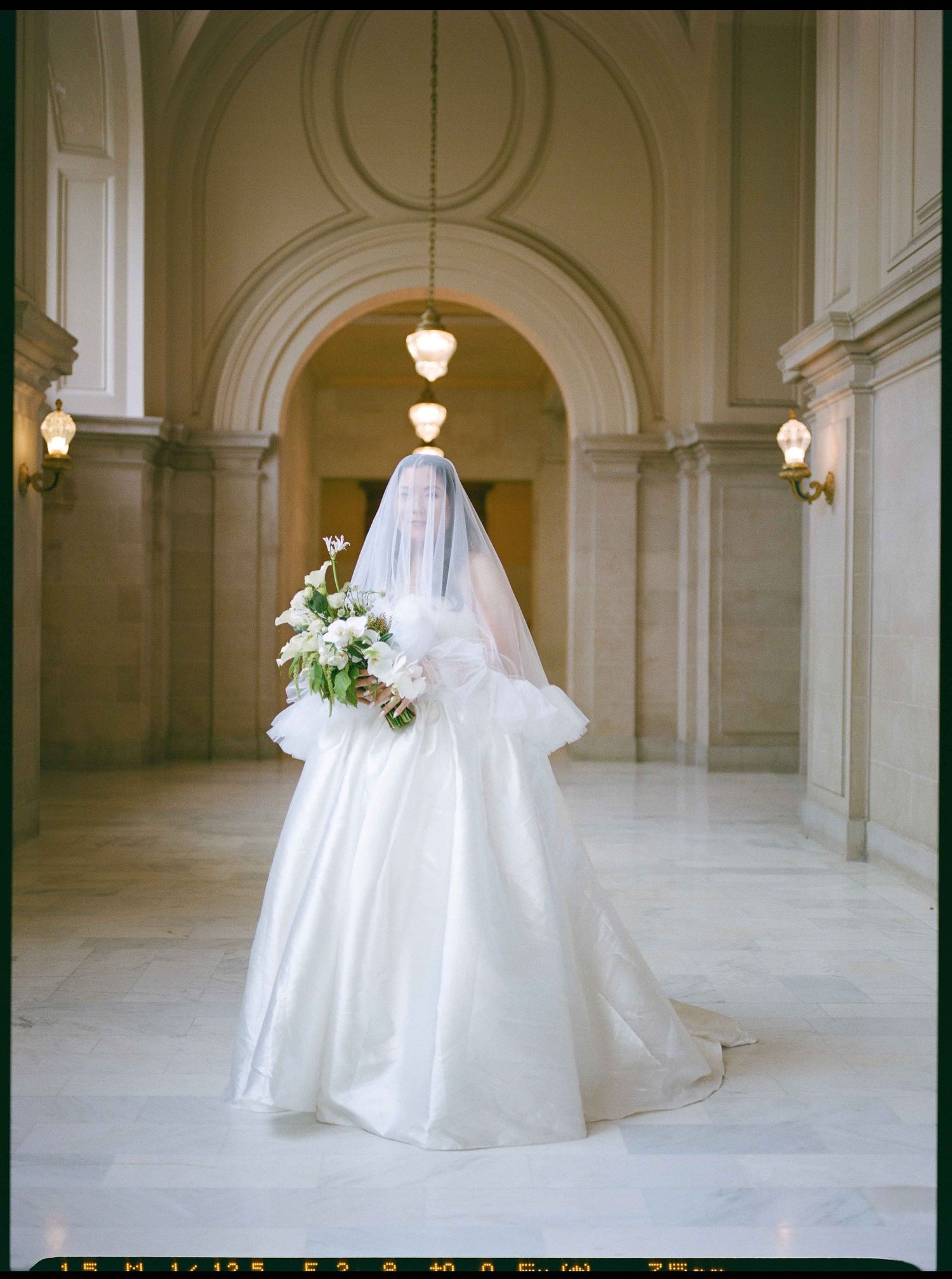 A bride in an elegant white wedding gown and long veil holds a bouquet while standing in a grand arched hallway.