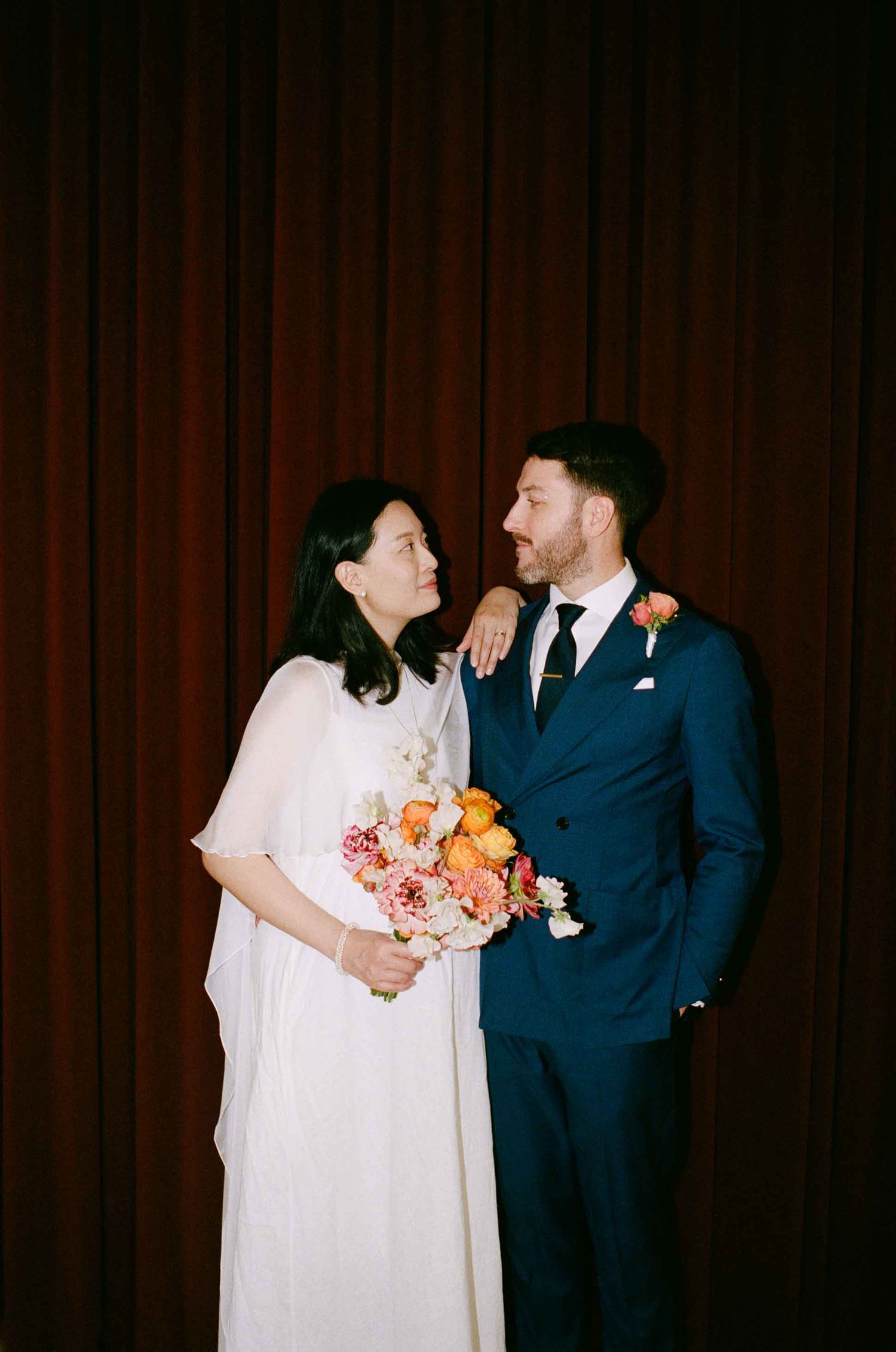 A couple poses together at their wedding, with the bride in white holding a colorful bouquet and groom in navy suit.