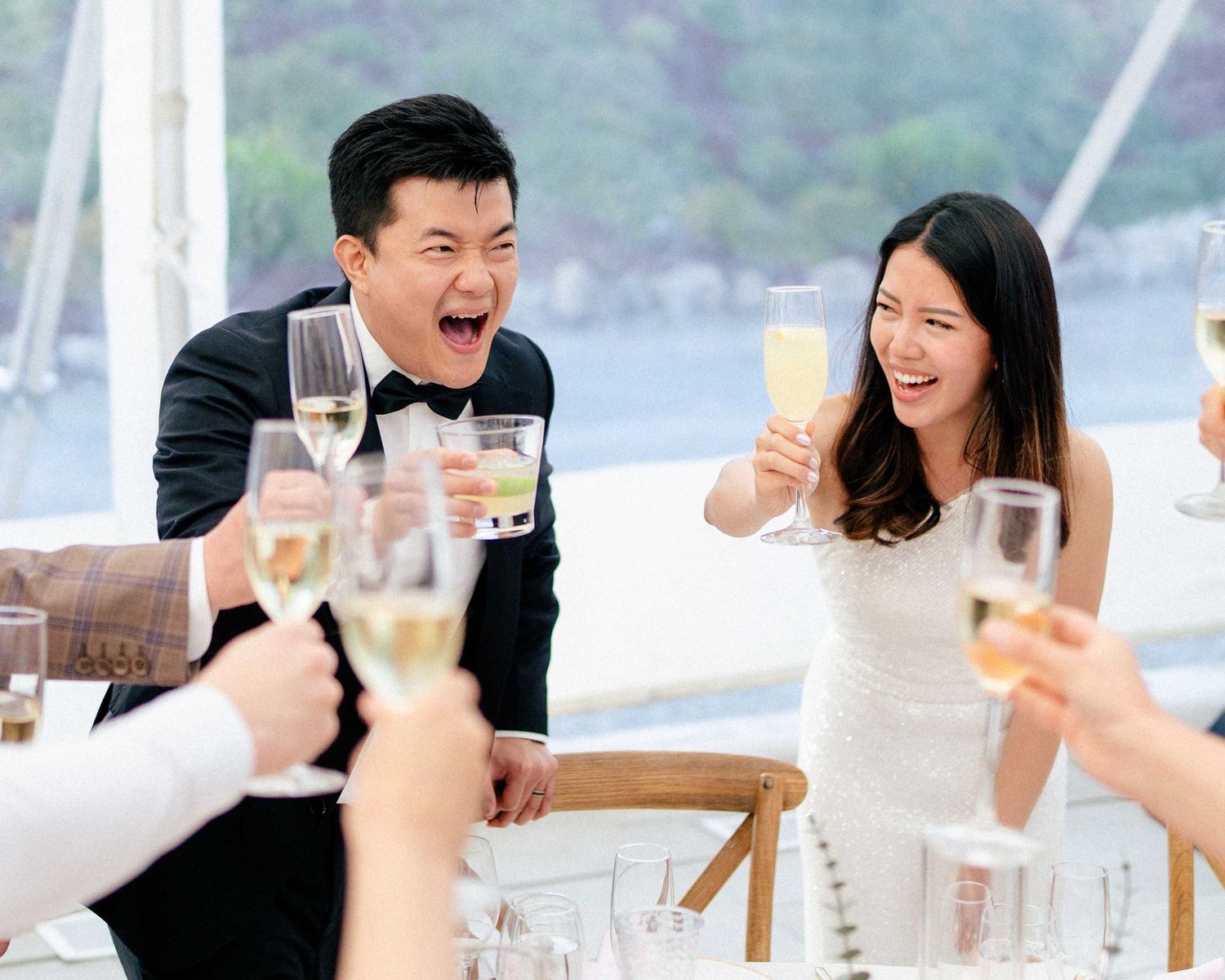 Wedding couple toasts champagne glasses with guests at an outdoor reception by the water.
