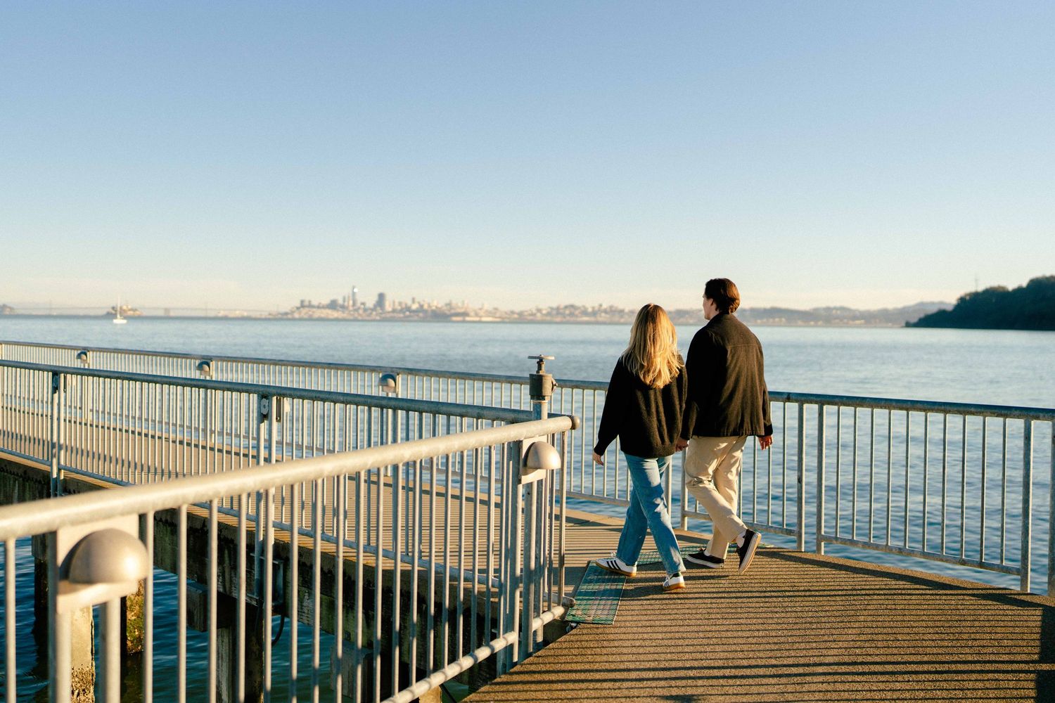 A couple walks along a waterfront pier with a city skyline visible in the distance.