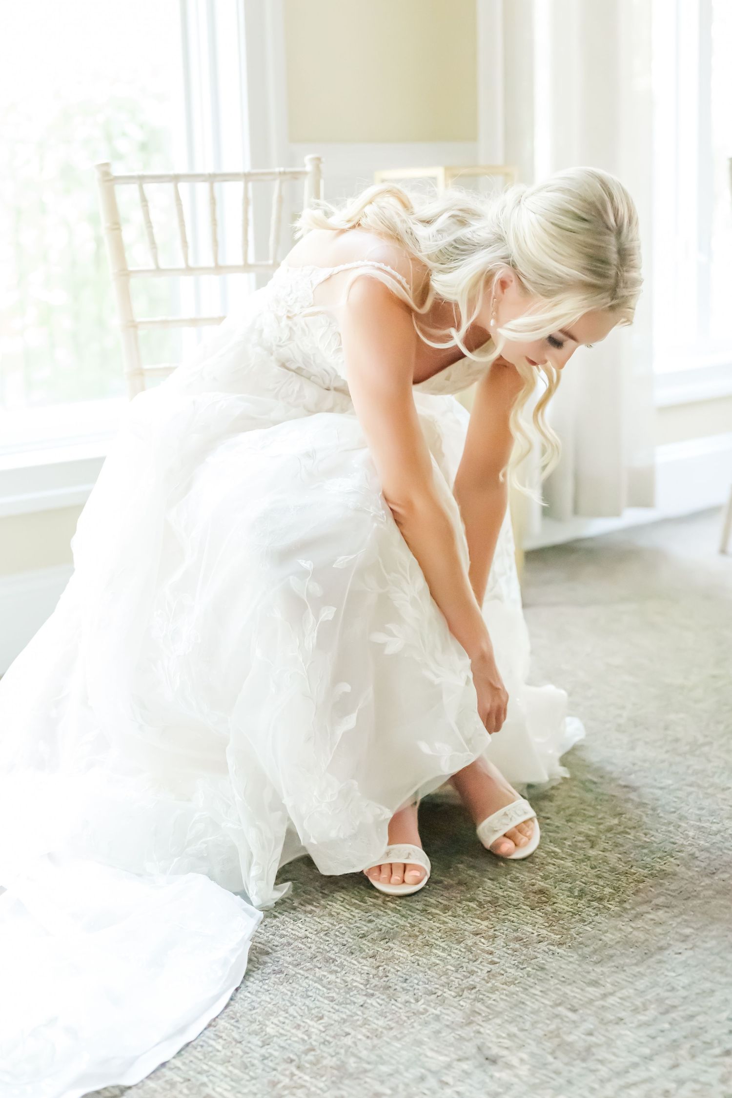 Bride in white wedding dress and heels bends down to adjust her shoe on a carpeted floor.