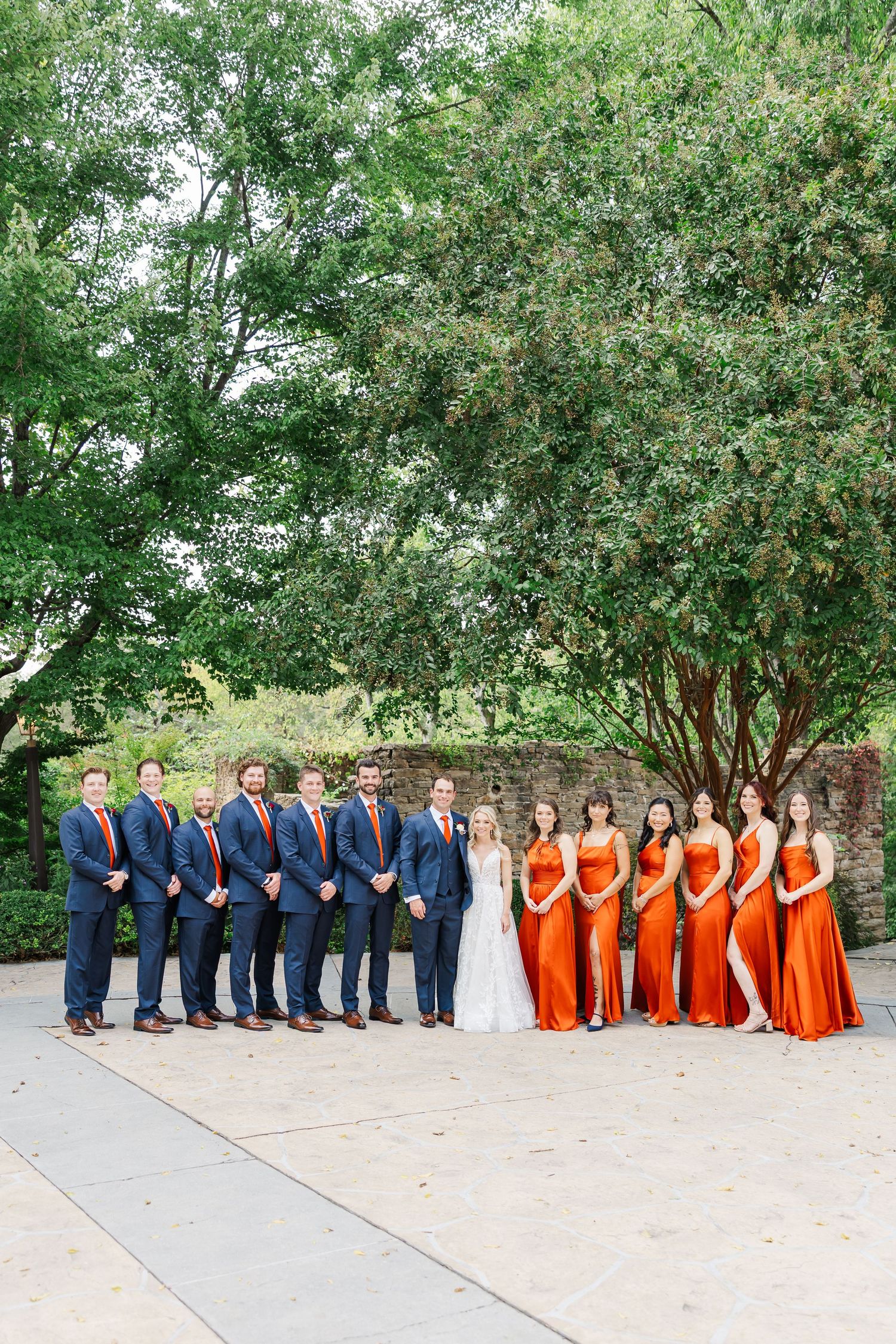 Wedding party in navy blue and orange attire poses under trees for formal group photos.