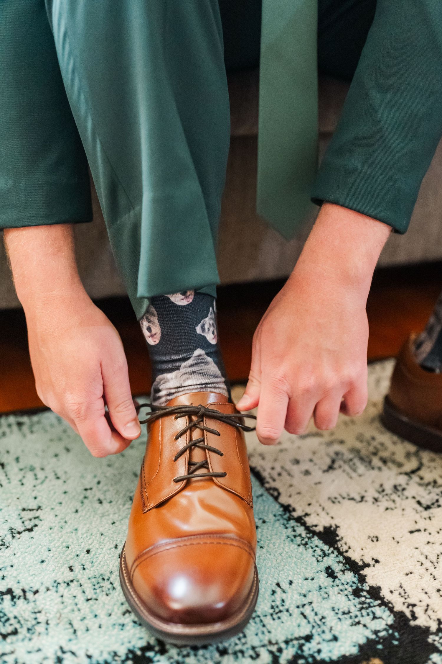 Person wearing green pants and fun patterned socks ties brown leather dress shoe on patterned rug.