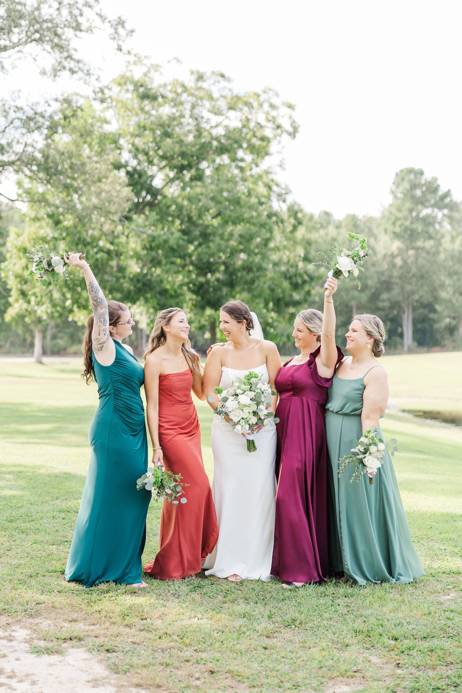 Wedding party in jewel-toned dresses stands together on a grassy lawn during an outdoor celebration.