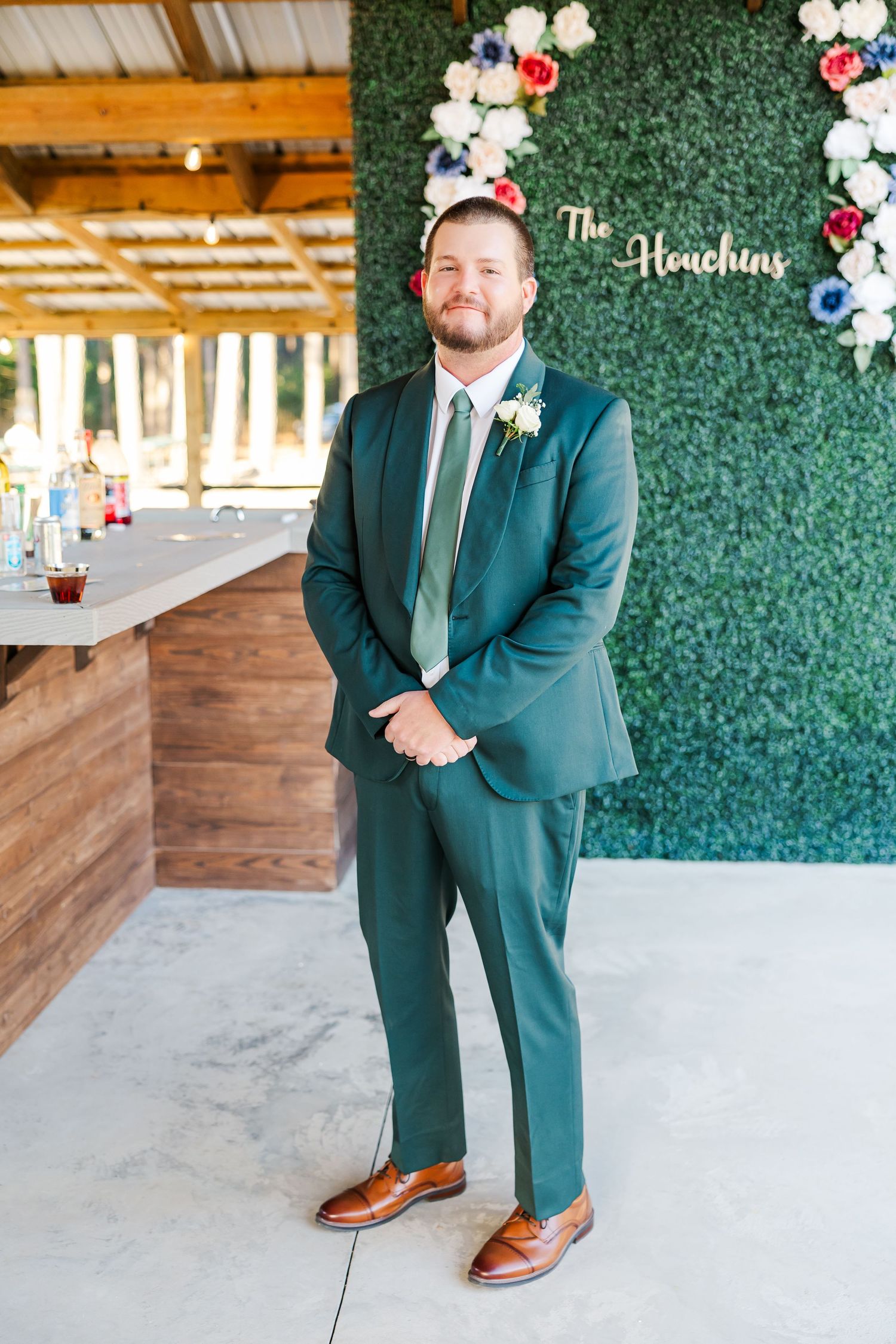 Groom in green suit with boutonniere poses before floral hedge wall at The Houchins wedding venue.