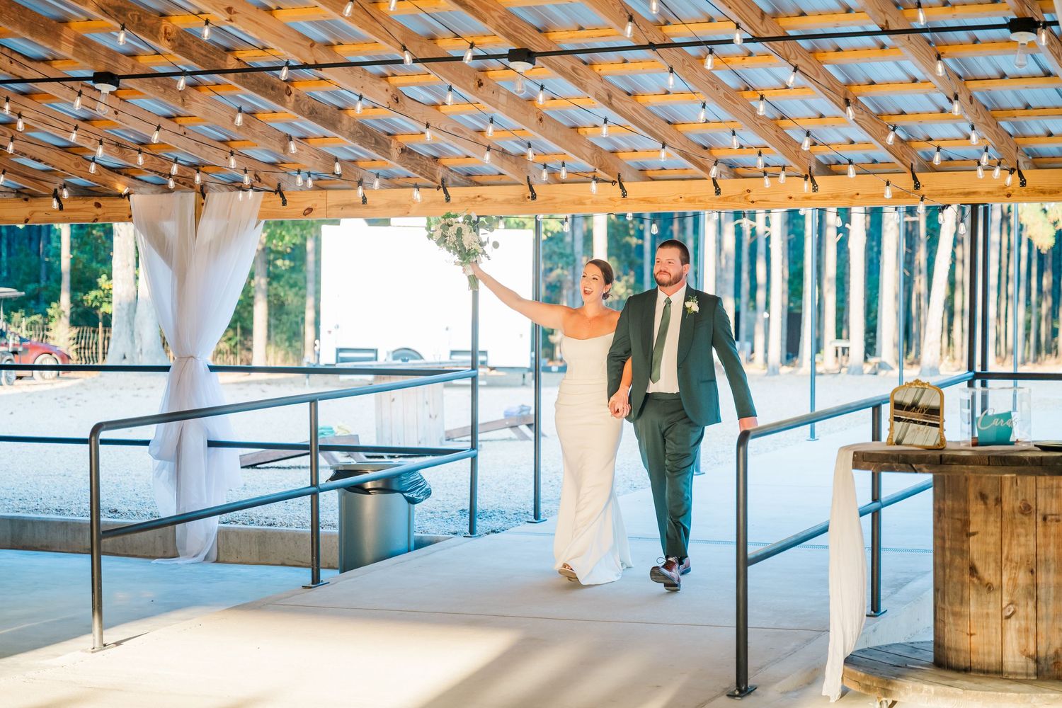 Bride and groom walking hand in hand under a rustic wooden pergola with string lights at their outdoor wedding venue.