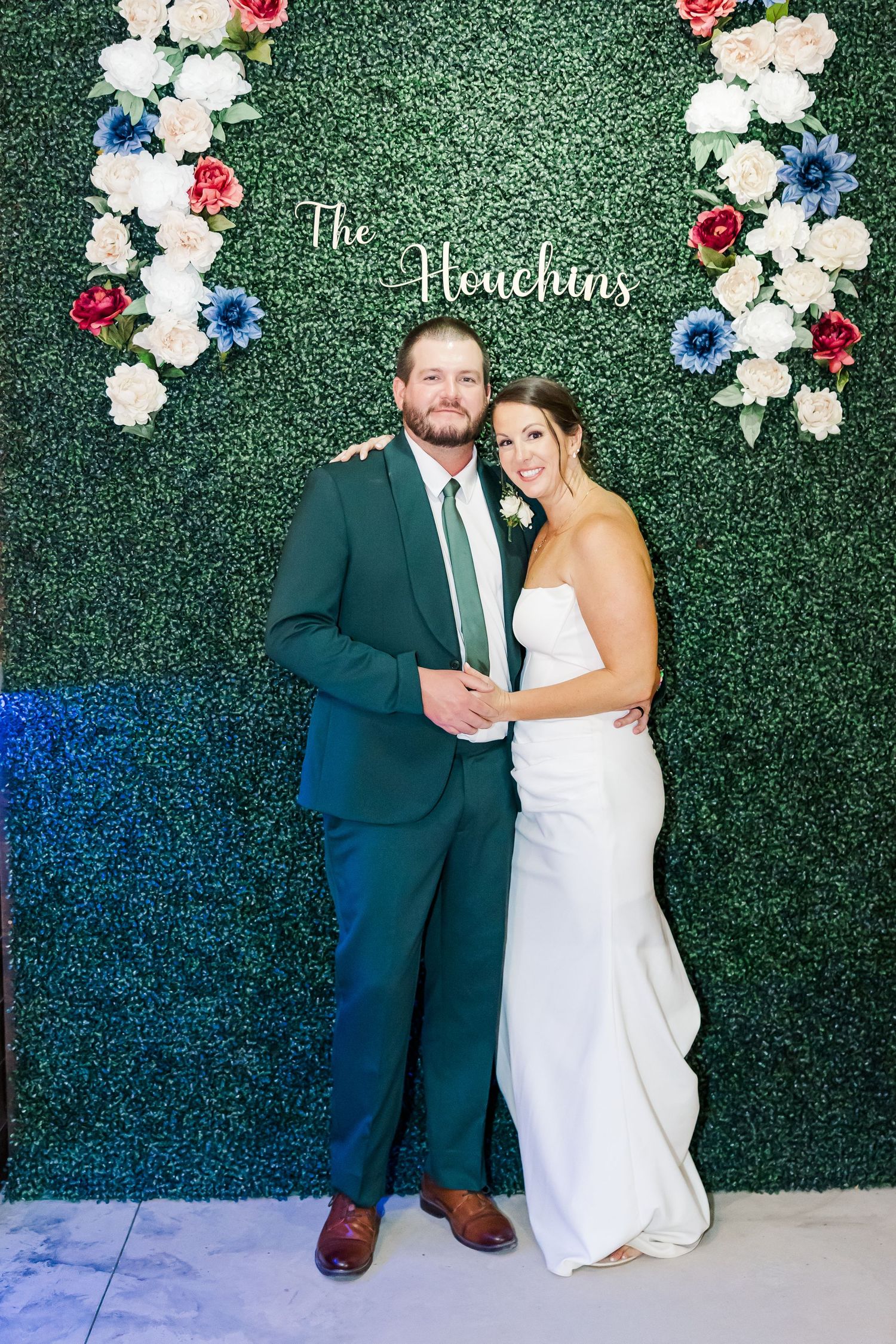 Bride and groom pose together in front of floral greenery wall backdrop reading The Houchins.