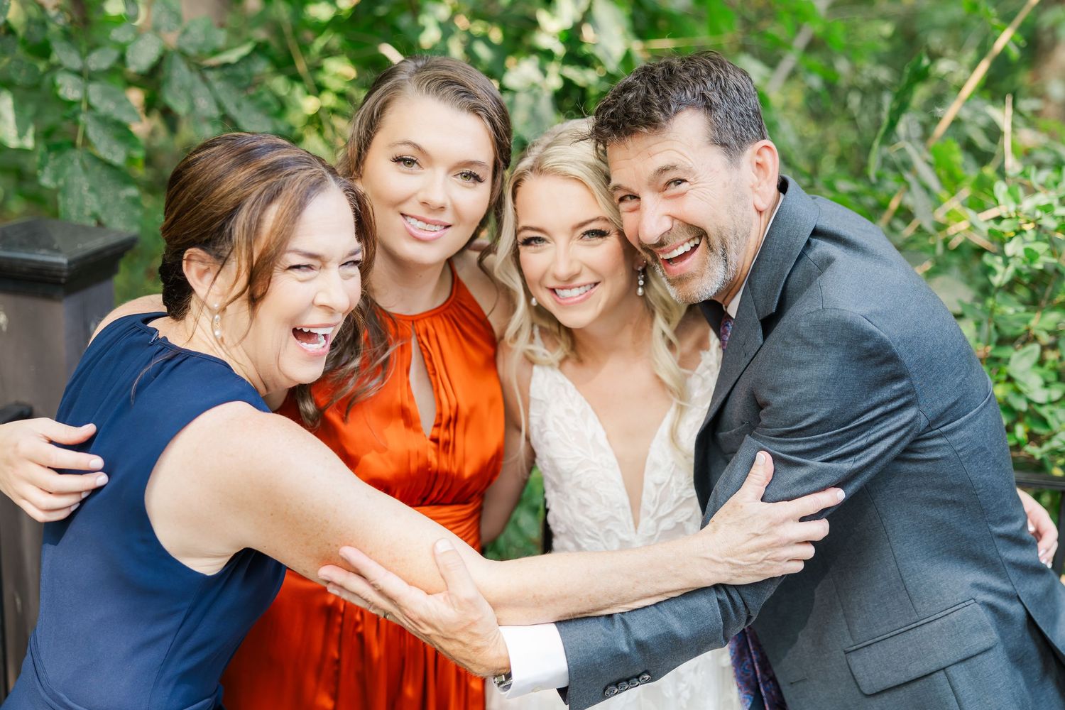 Wedding party laughs together while posing in a lush green garden setting during a joyful celebration.